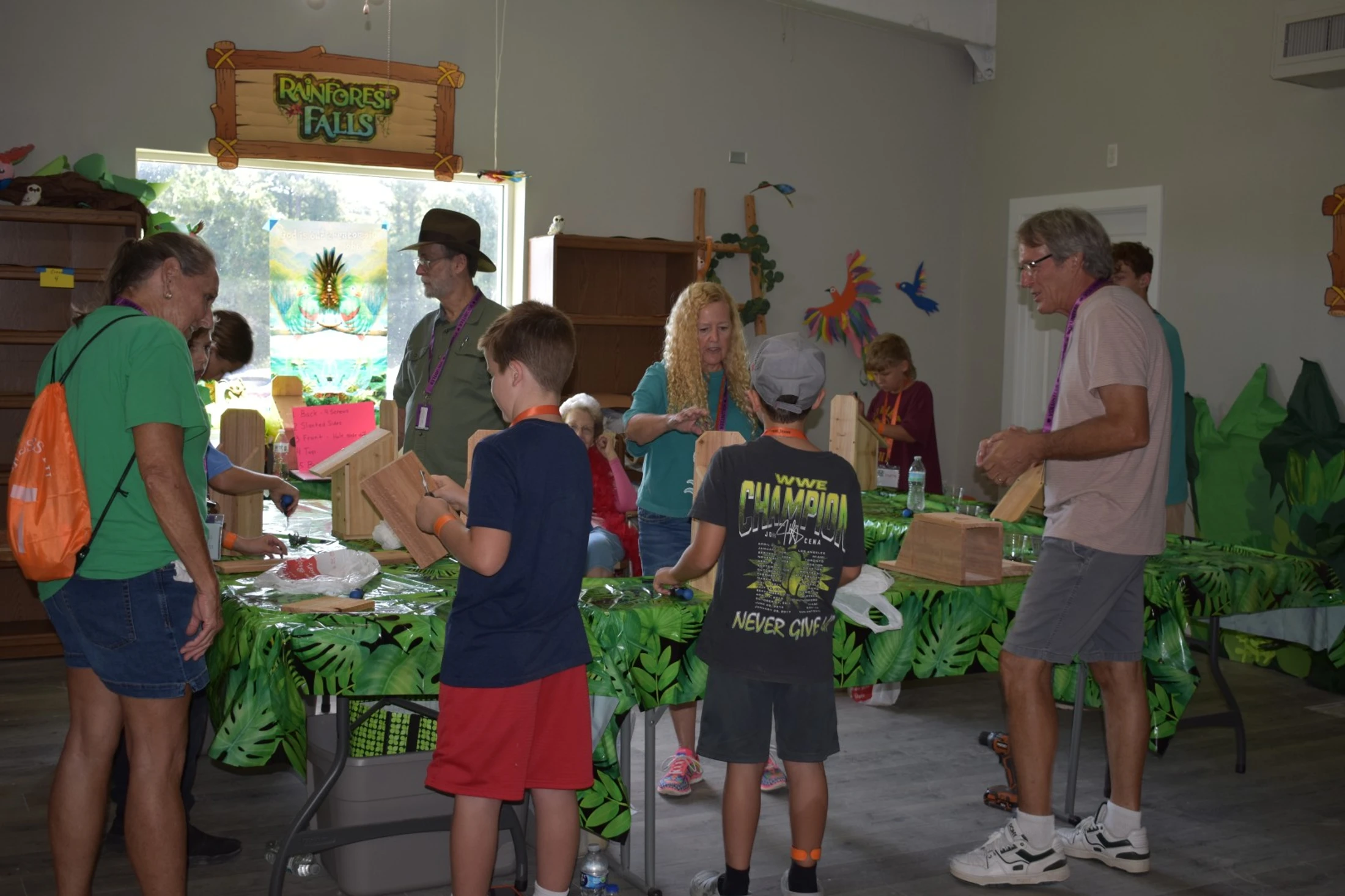 Adults and children gather around tables with leafy tablecloths, building wooden crafts in a room decorated with a 'Rainforest Falls' sign.