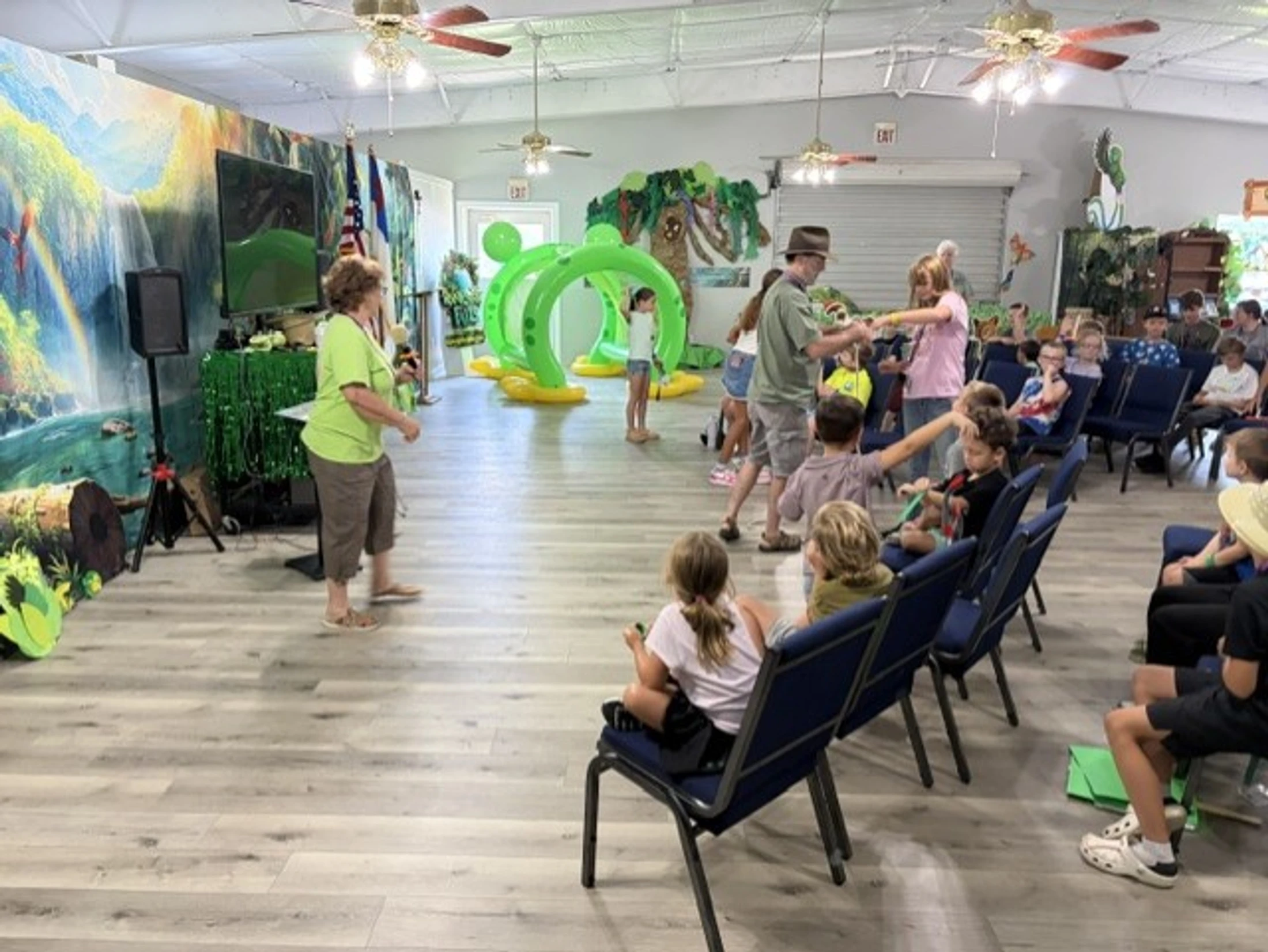 A large room decorated for a kids' jungle-themed event. Children are seated on chairs or playing near inflatables, with adults supervising the activities.