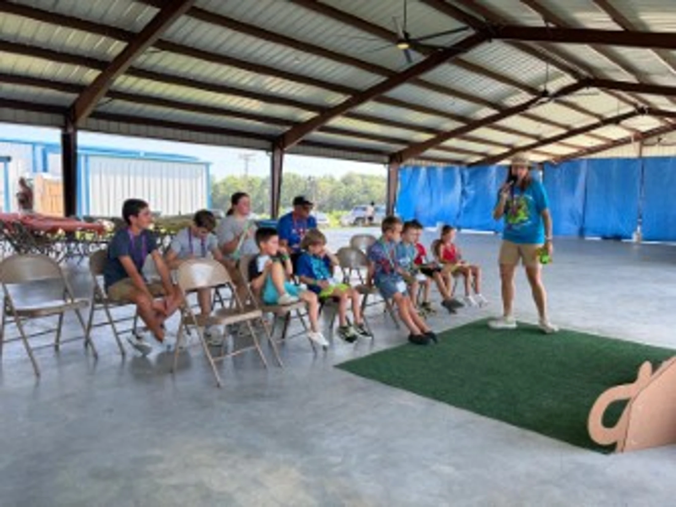 A group of children and a few adults sit on folding chairs in an open-air pavilion, listening to a woman with a microphone giving a presentation.