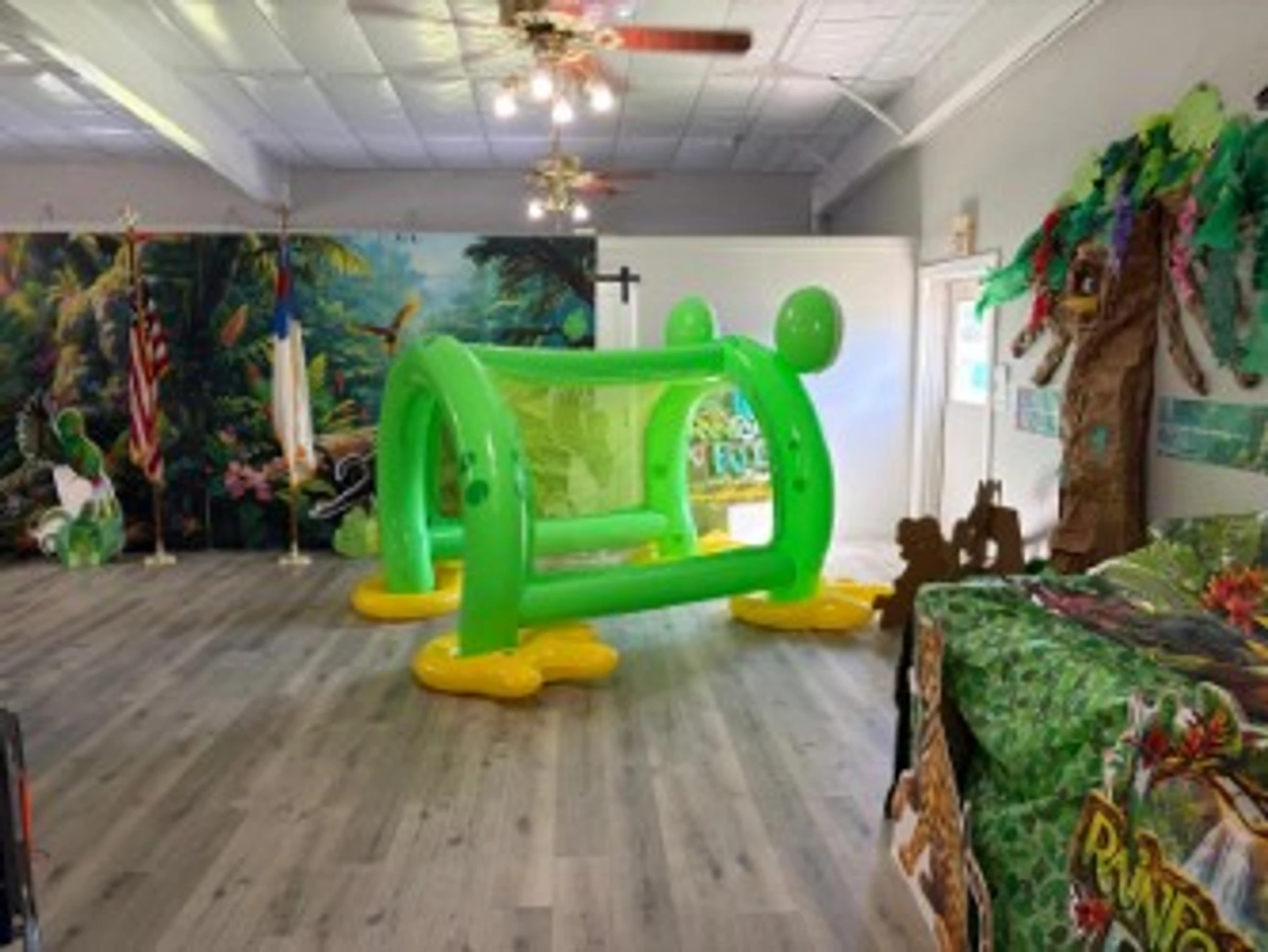 An indoor room decorated with a jungle theme, featuring a large, green inflatable frog soccer goal in the center on a gray wood-look floor.