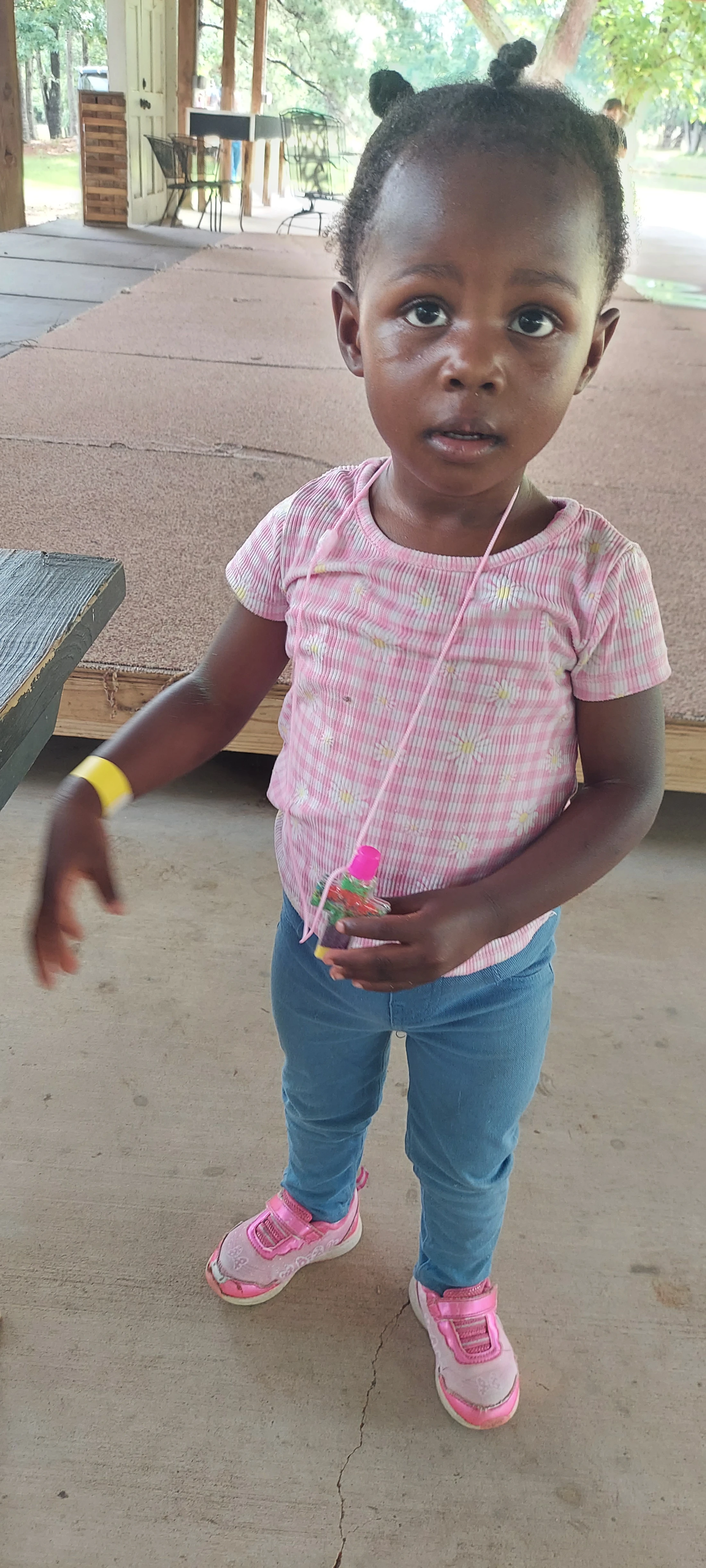 A young Black girl with her hair in bantu knots stands on a covered patio. She wears a pink shirt, jeans, and pink shoes and looks directly at the camera.