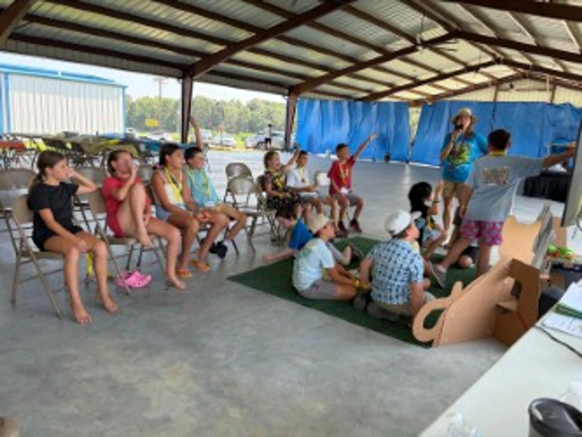 A group of children sits under an outdoor pavilion listening to an instructor. Some children have their hands raised, eager to participate in the activity.