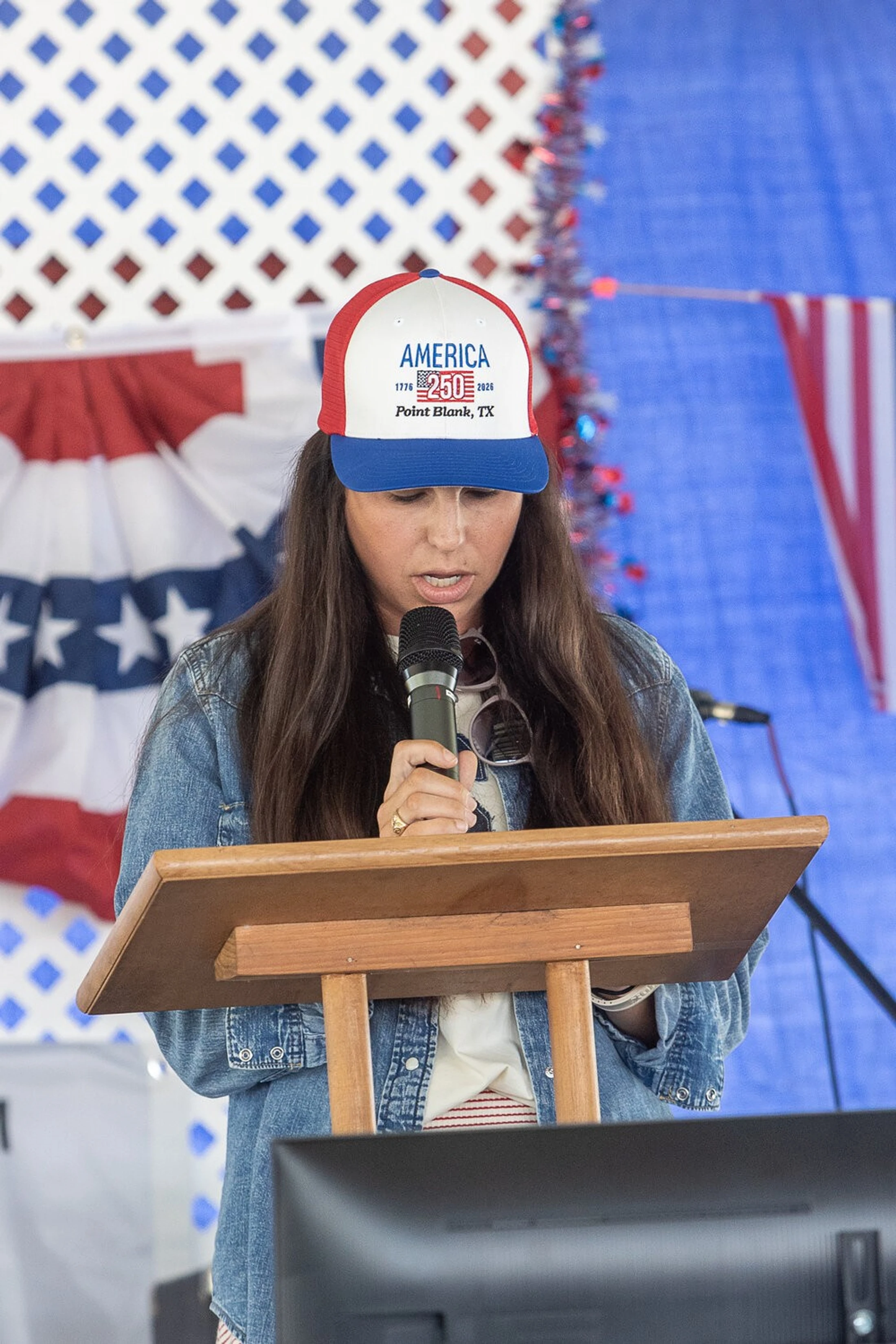A woman wearing a red, white, and blue 'America 250' hat and a denim jacket speaks into a microphone at a wooden podium with patriotic decorations behind her.