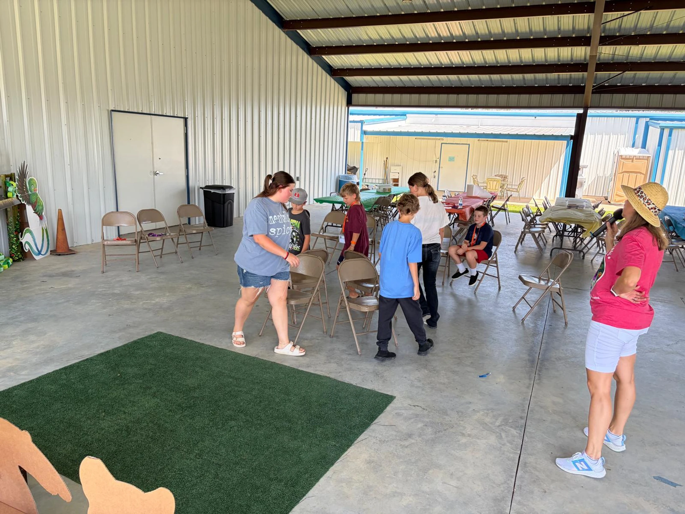 A group of children and adults in a large, open-sided pavilion arrange folding chairs on a concrete floor, preparing for a group game or activity.