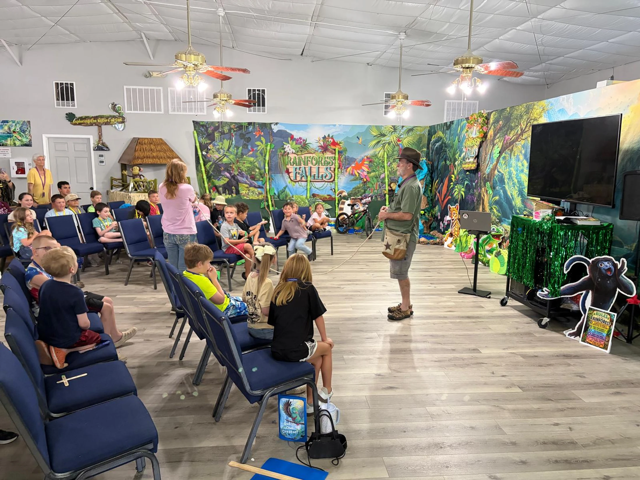 A group of children sit on blue chairs watching a man in an explorer costume give a presentation in a room decorated with a colorful rainforest theme.
