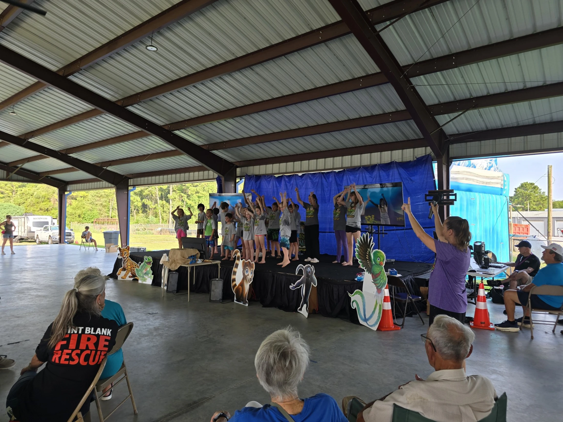 A group of children performs on a stage under a large pavilion for an audience of adults sitting in folding chairs. A woman leads the kids from the side.