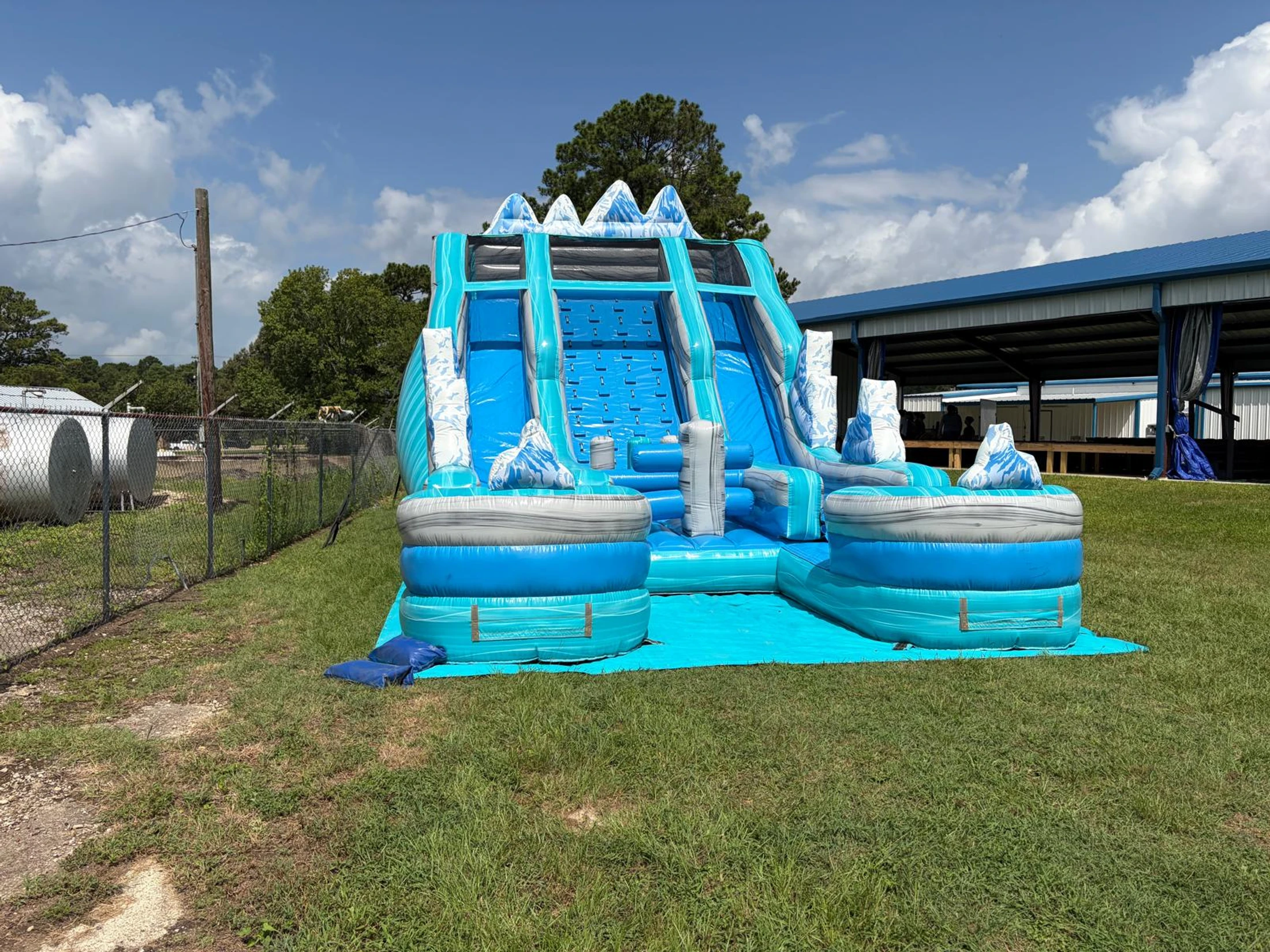 A large, blue and white inflatable water slide with an arctic mountain theme sits on a grassy field under a partly cloudy sky, ready for an event.
