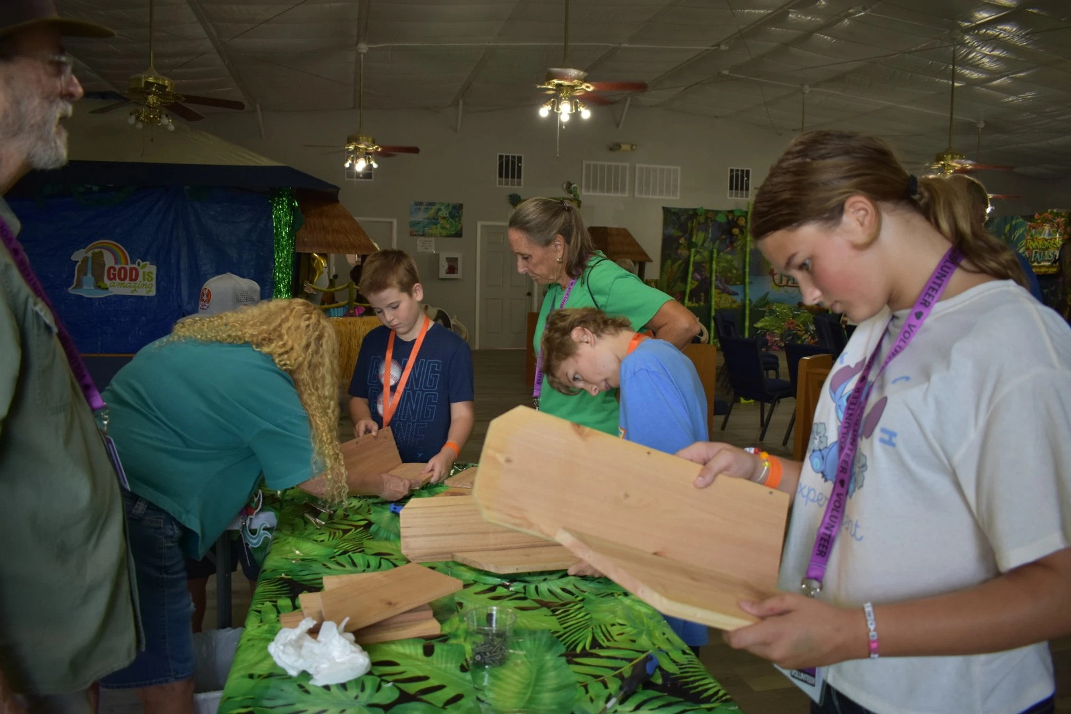 A group of kids and adult volunteers gather around a table with a leaf-patterned cover, focused on assembling small wooden craft projects indoors.