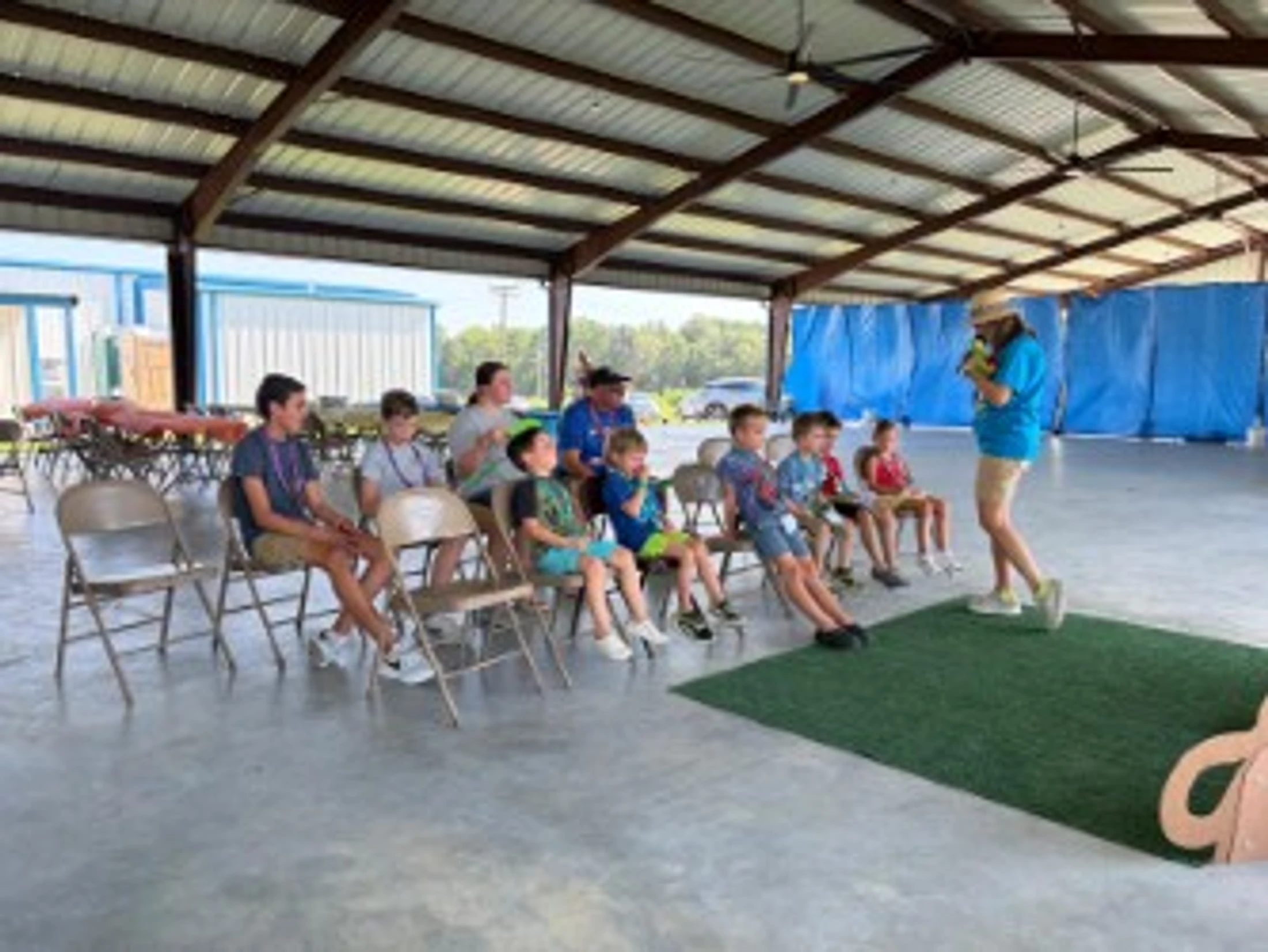 A group of children sits on chairs under a large open-air pavilion, listening to a woman who is standing and giving a presentation.