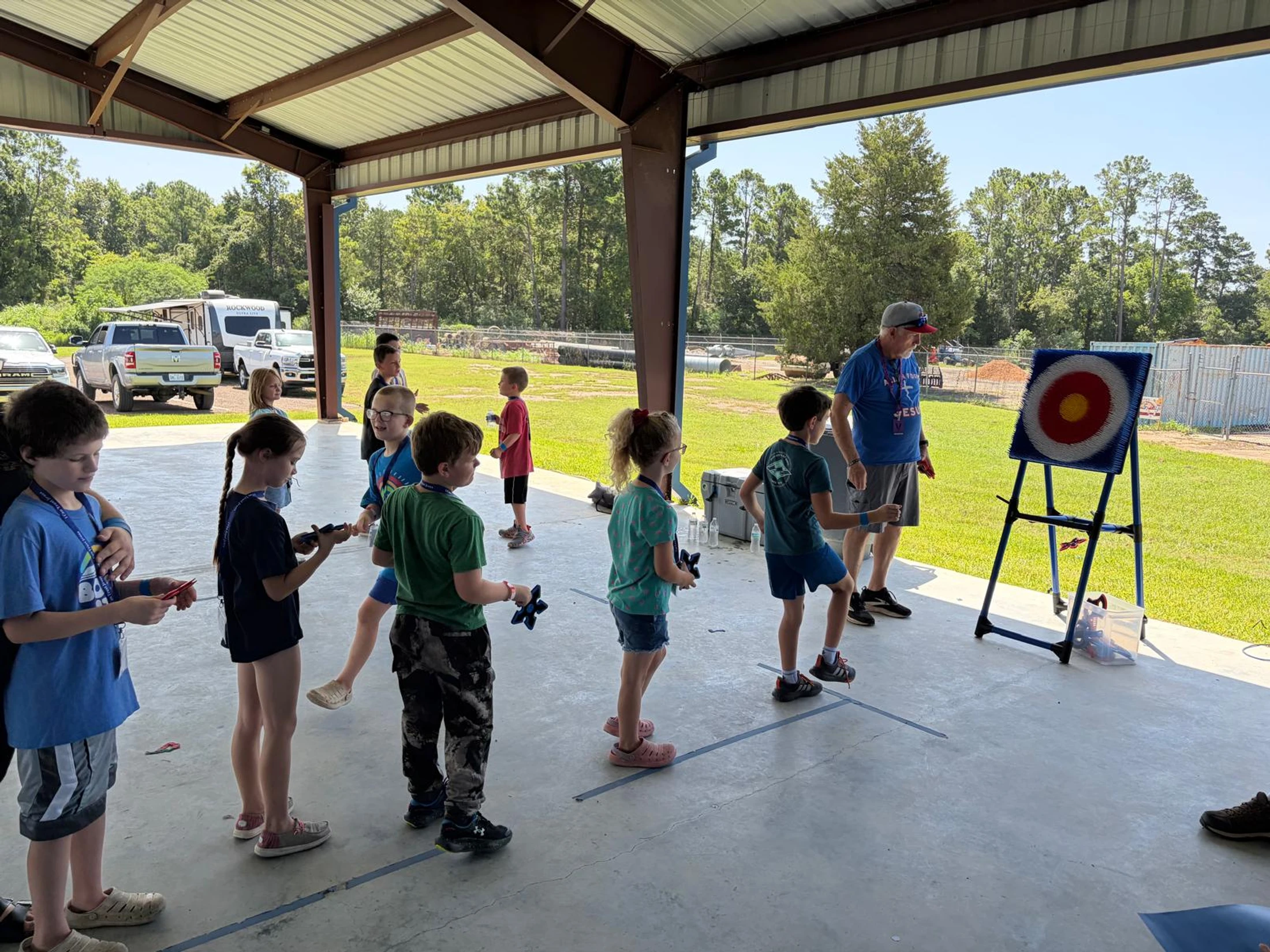 A group of children line up under a covered pavilion to throw star-shaped projectiles at a red and yellow target, supervised by an adult man.