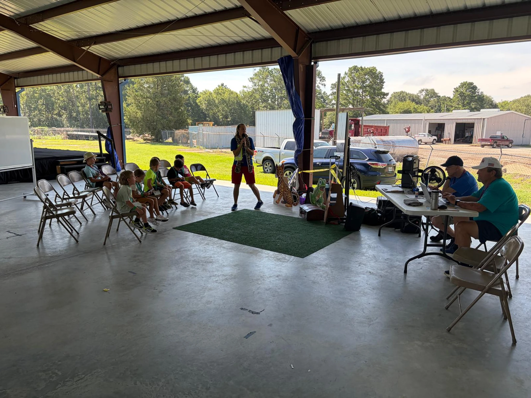 A man gives a presentation to a group of children seated on folding chairs under a large, open-air pavilion, with two men at a table nearby.