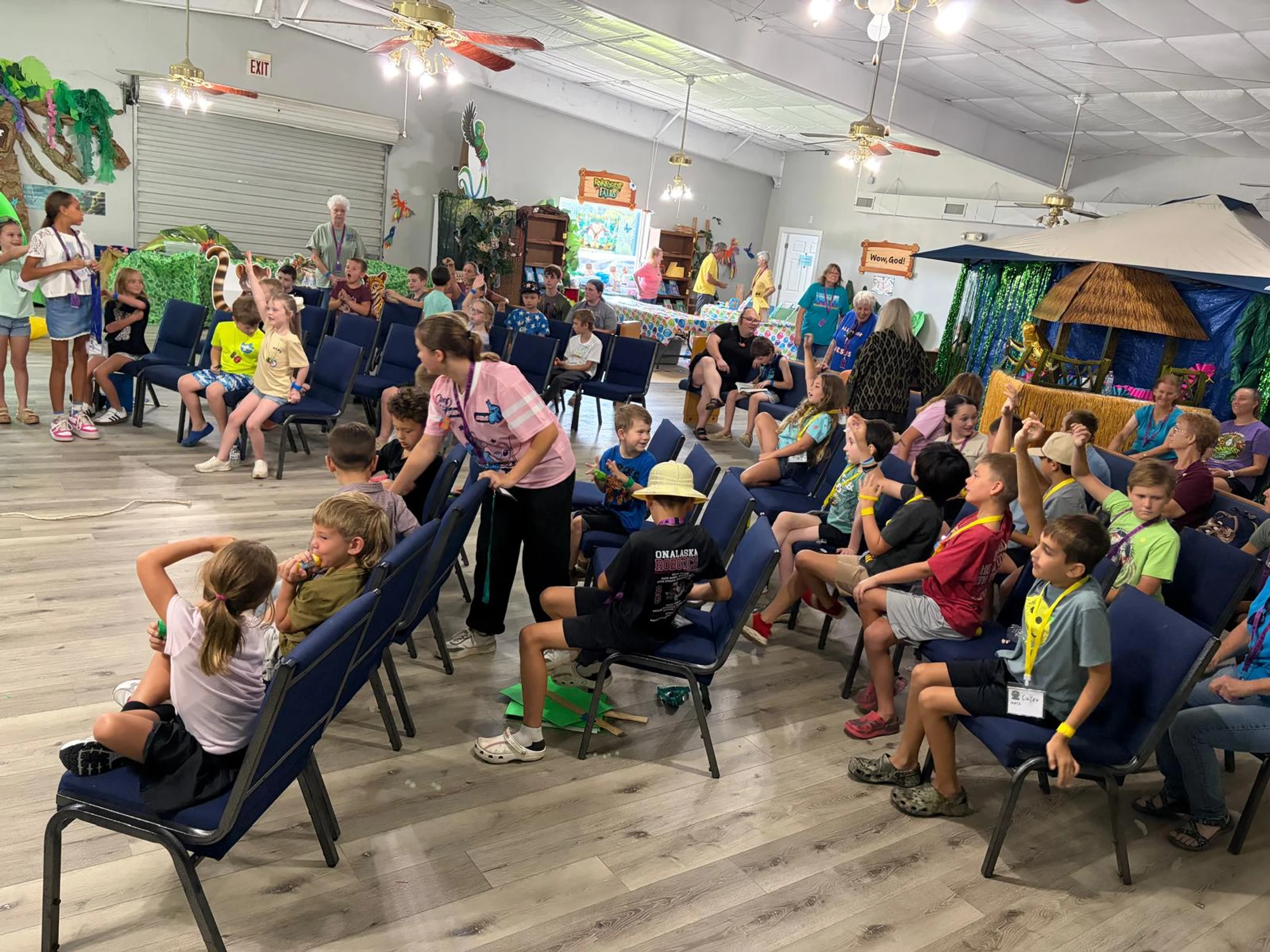 A large group of children and adults gathered in a brightly lit room for a jungle-themed event, with many children sitting in chairs and raising their hands.