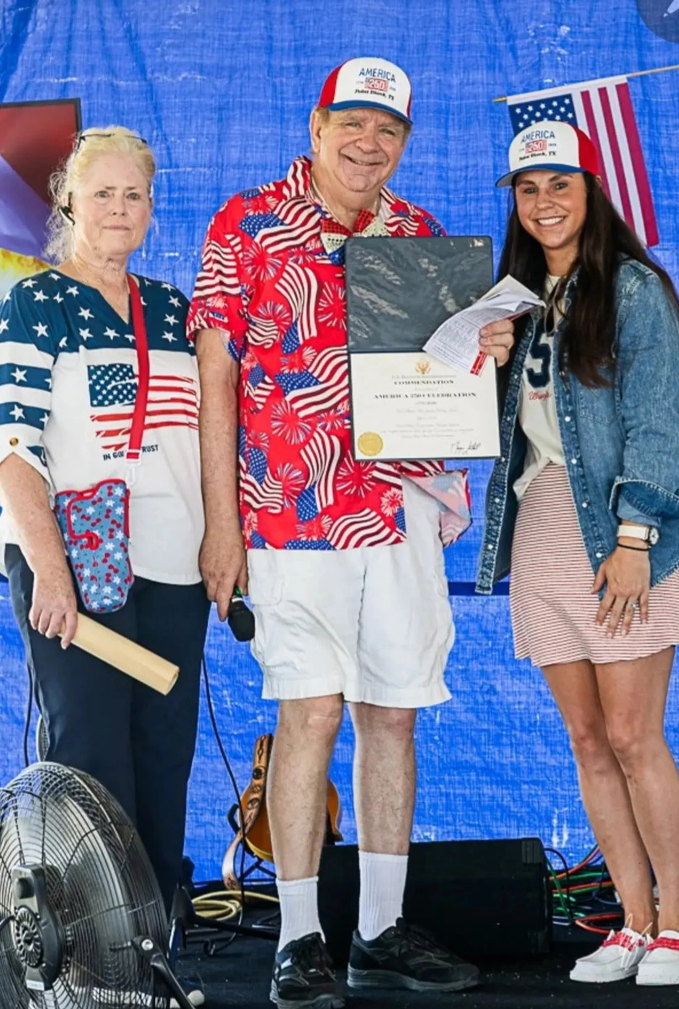 A man and two women in patriotic American flag-themed outfits pose for a photo on a stage. The man in the center holds a certificate of commendation.