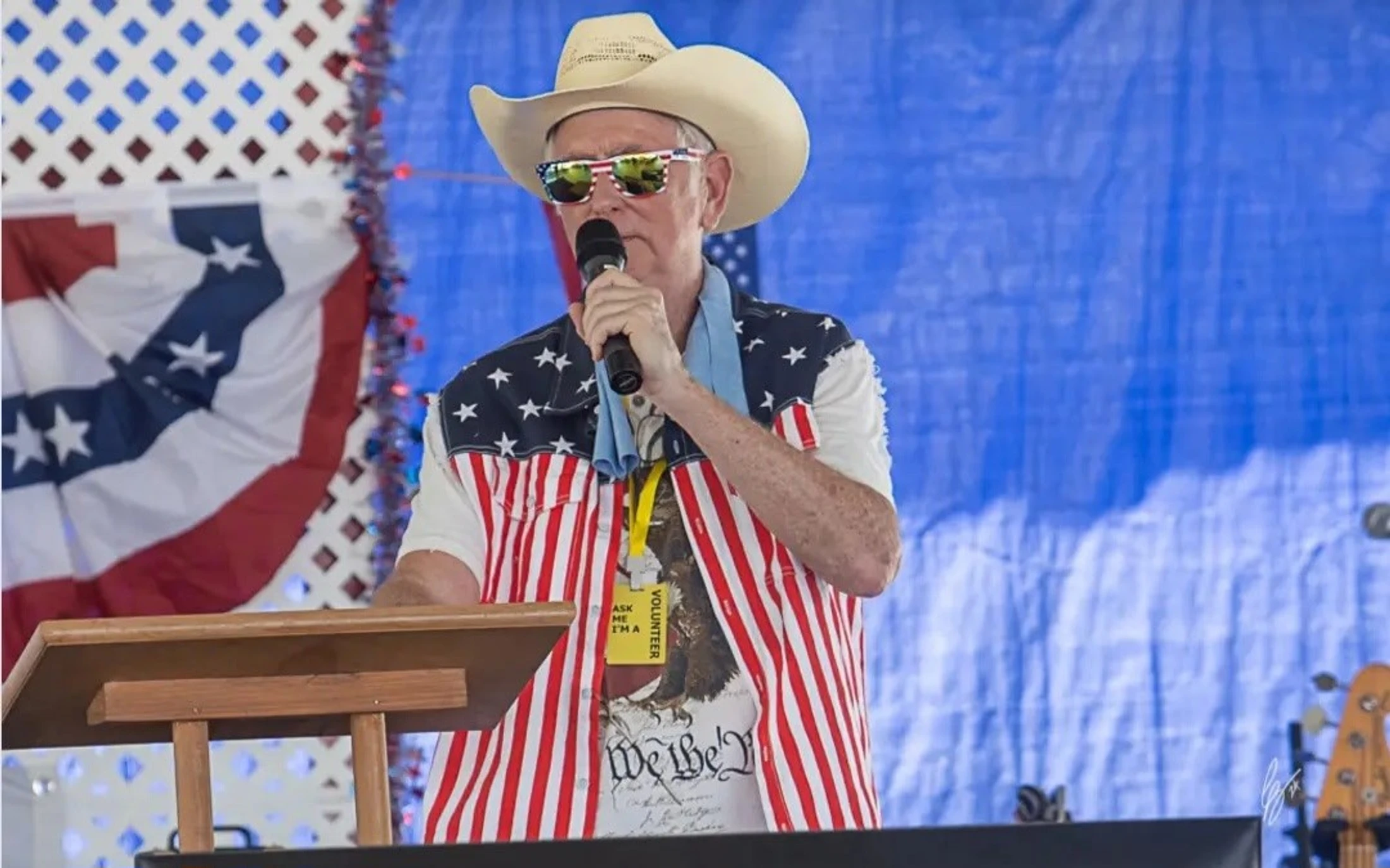 A man in a cowboy hat, American flag sunglasses, and a stars and stripes vest speaks into a microphone behind a podium with patriotic decorations behind him.
