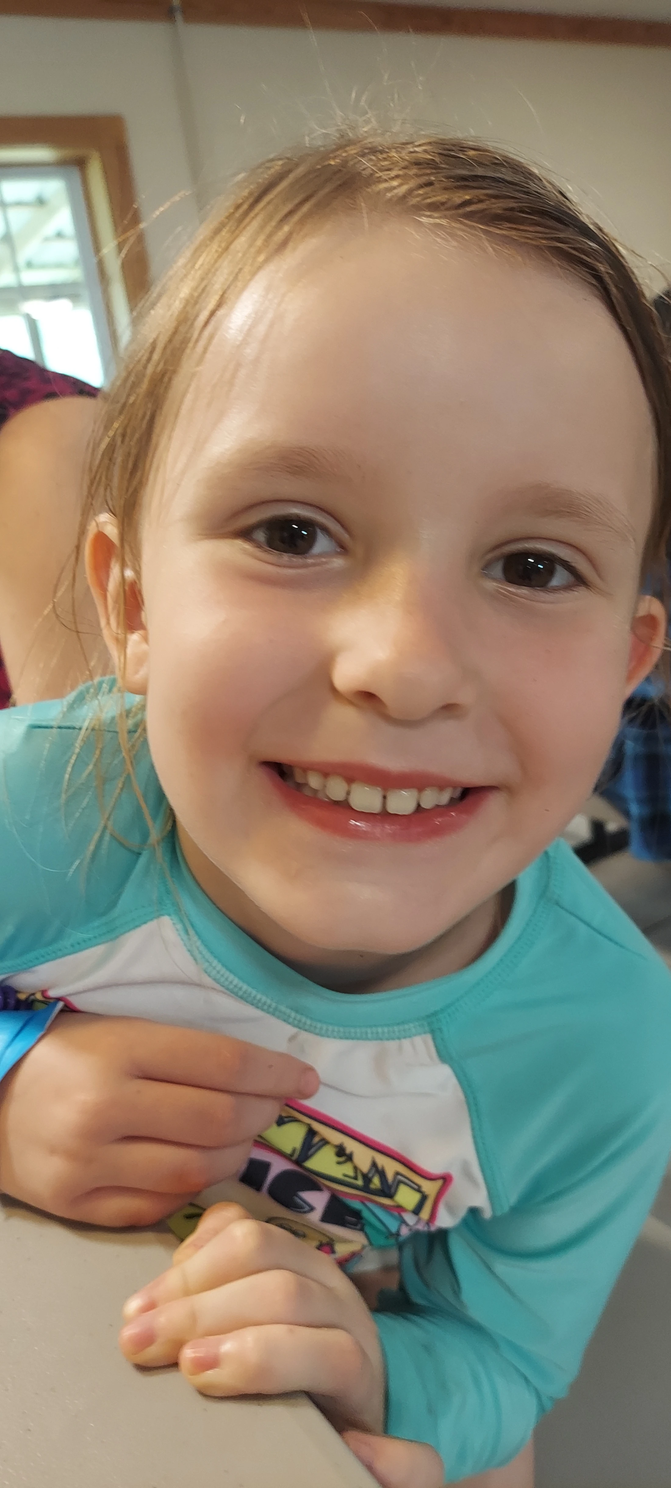 A close-up portrait of a young girl with light brown hair and big brown eyes, smiling happily at the camera while wearing a turquoise swim shirt.