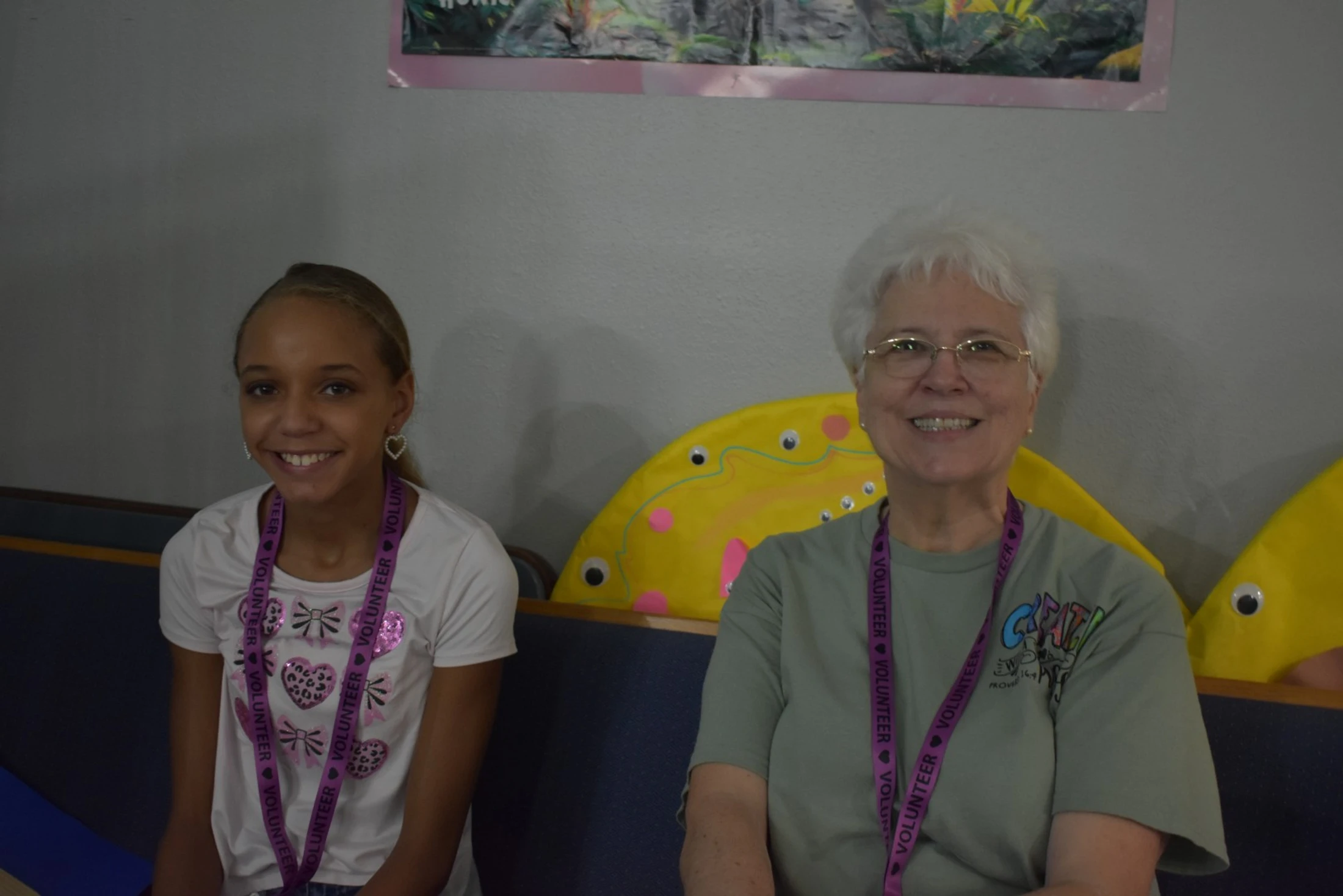A young girl and an older woman sit next to each other on a blue bench, smiling. Both are wearing purple lanyards that say 'VOLUNTEER'.