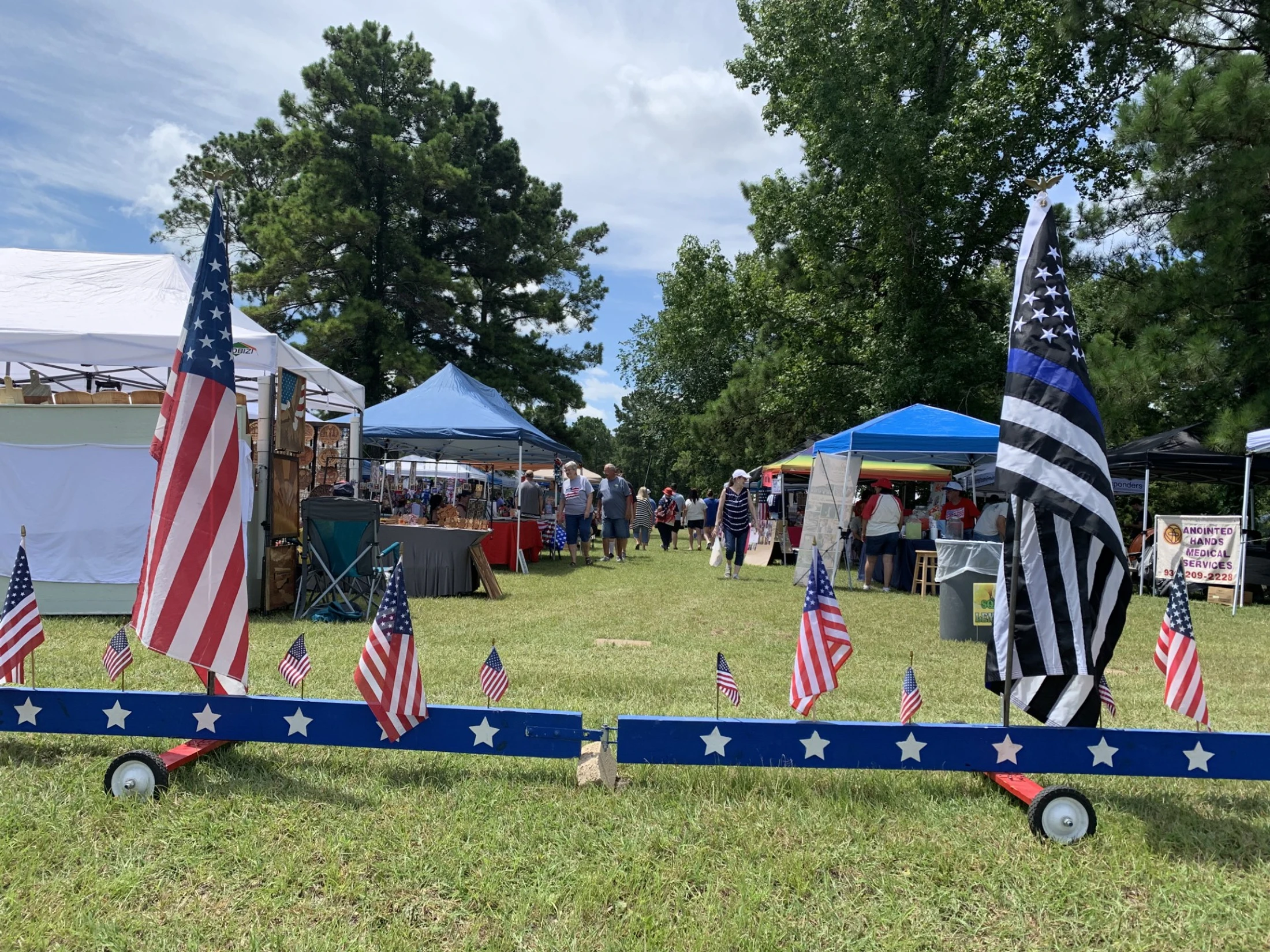An outdoor market with people browsing stalls. In the foreground, an American flag and a Thin Blue Line flag are displayed on a star-spangled wooden beam.