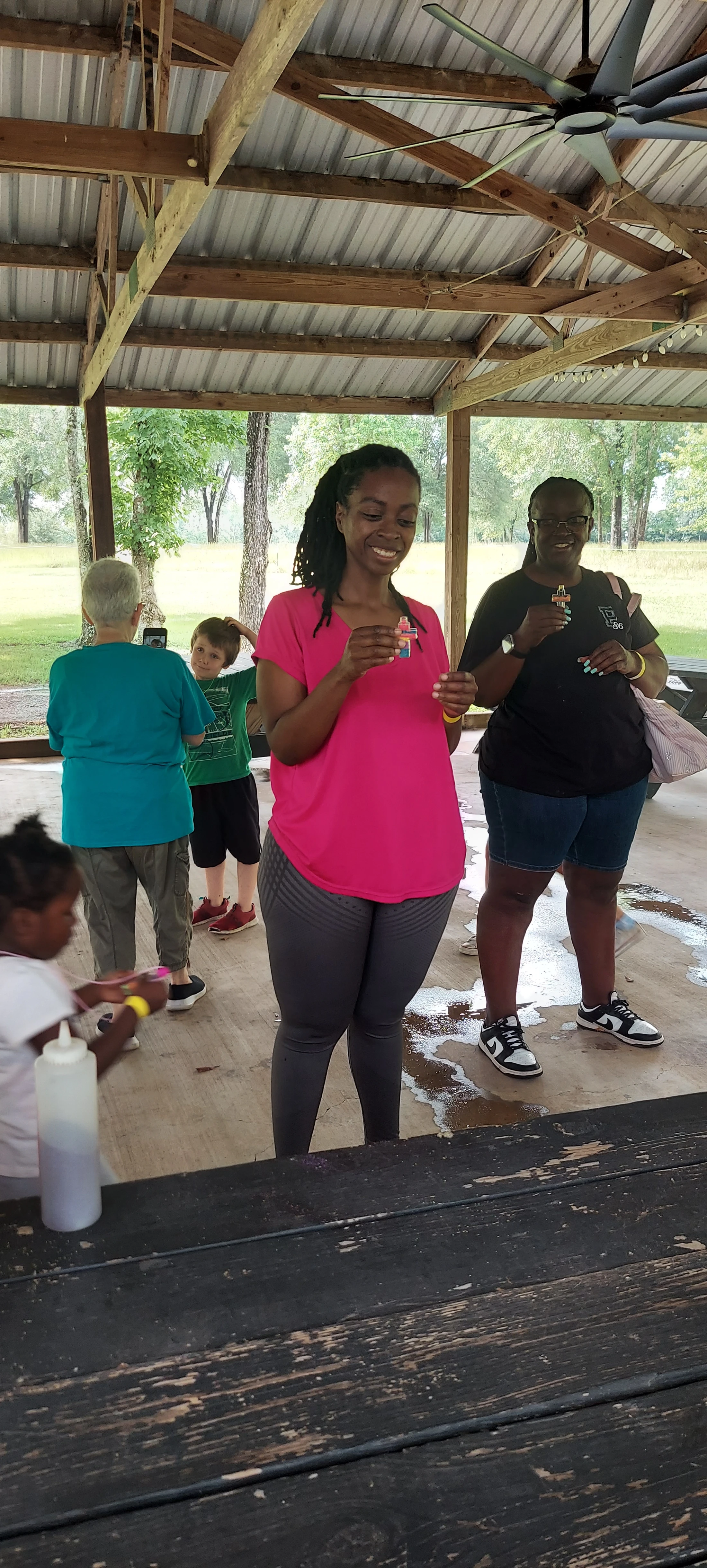A diverse group of adults and children stand under a park pavilion. Two women in the center smile as they hold up small, colorful handmade crosses.