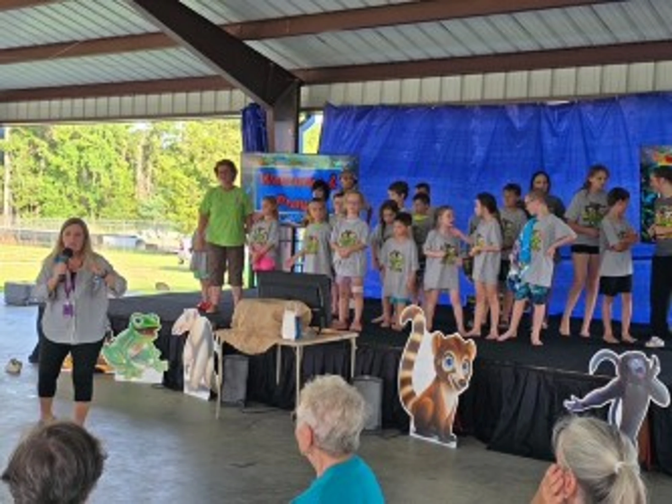A woman with a microphone speaks to an audience as a group of children and another adult stand on a stage decorated with cartoon animal cutouts.
