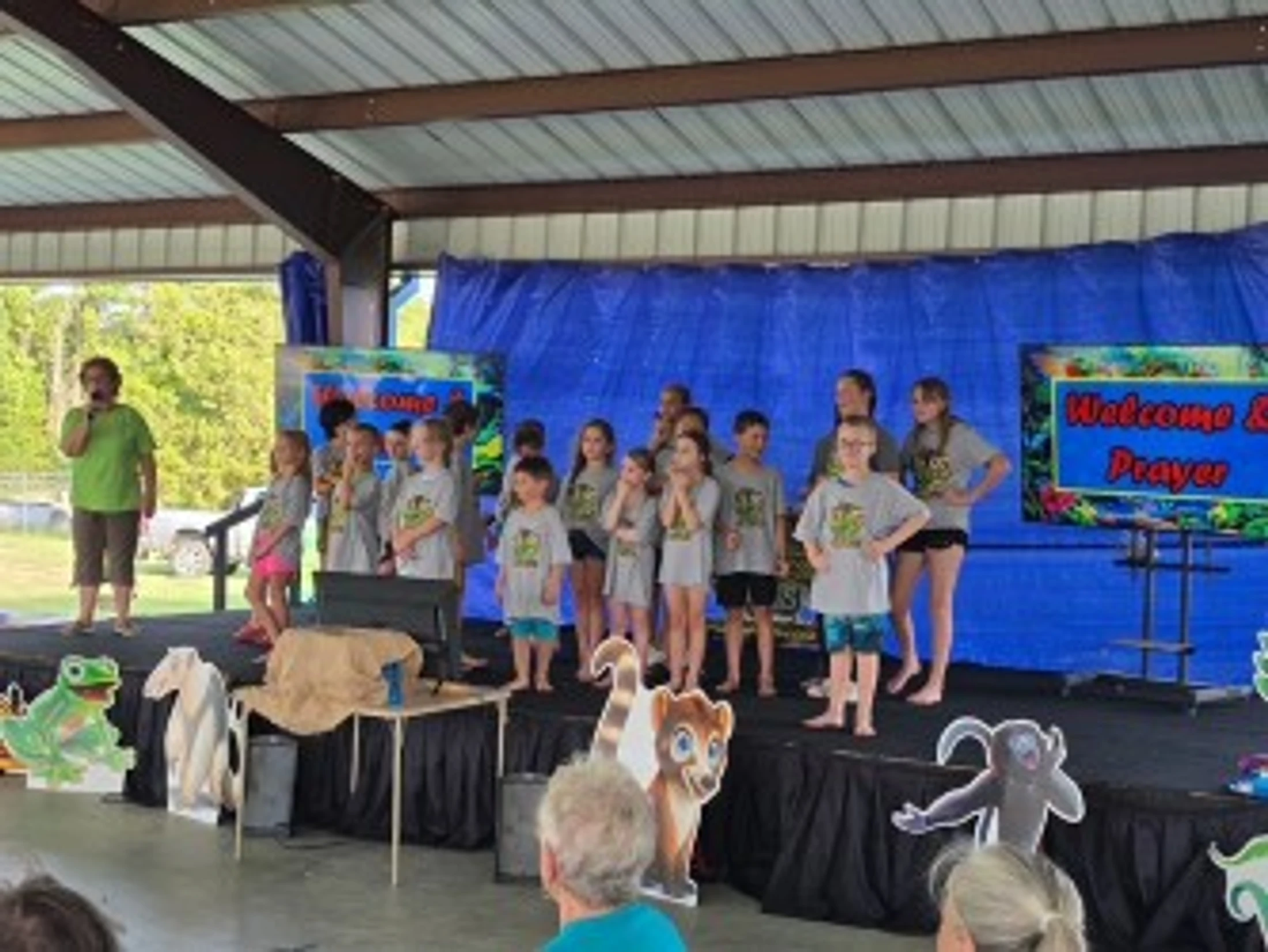 A group of children and an adult leader stand on a decorated stage under a pavilion, appearing to sing or perform for an audience at an outdoor event.