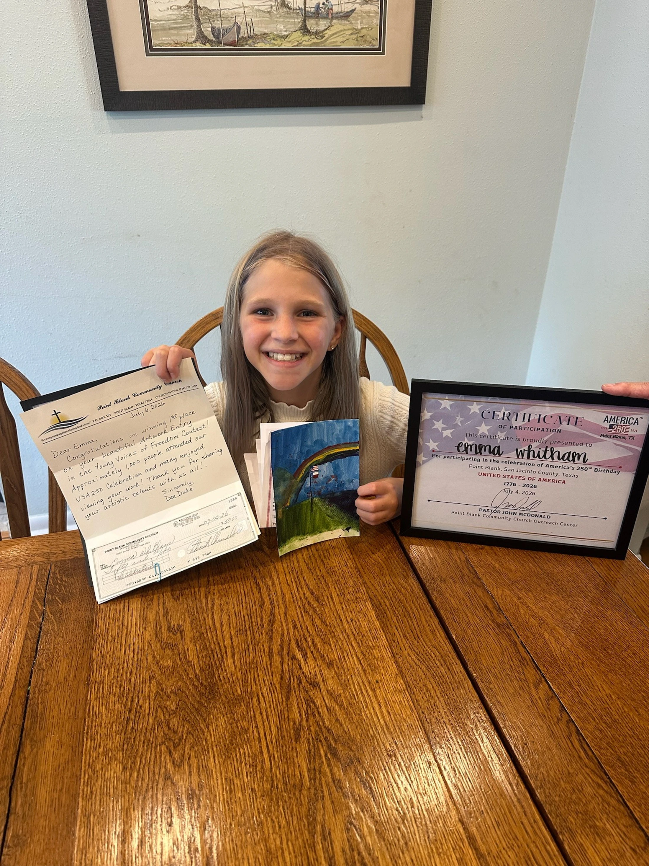 A smiling young girl sits at a wooden table showing her 1st place letter, prize check, painting, and framed certificate from an art contest.