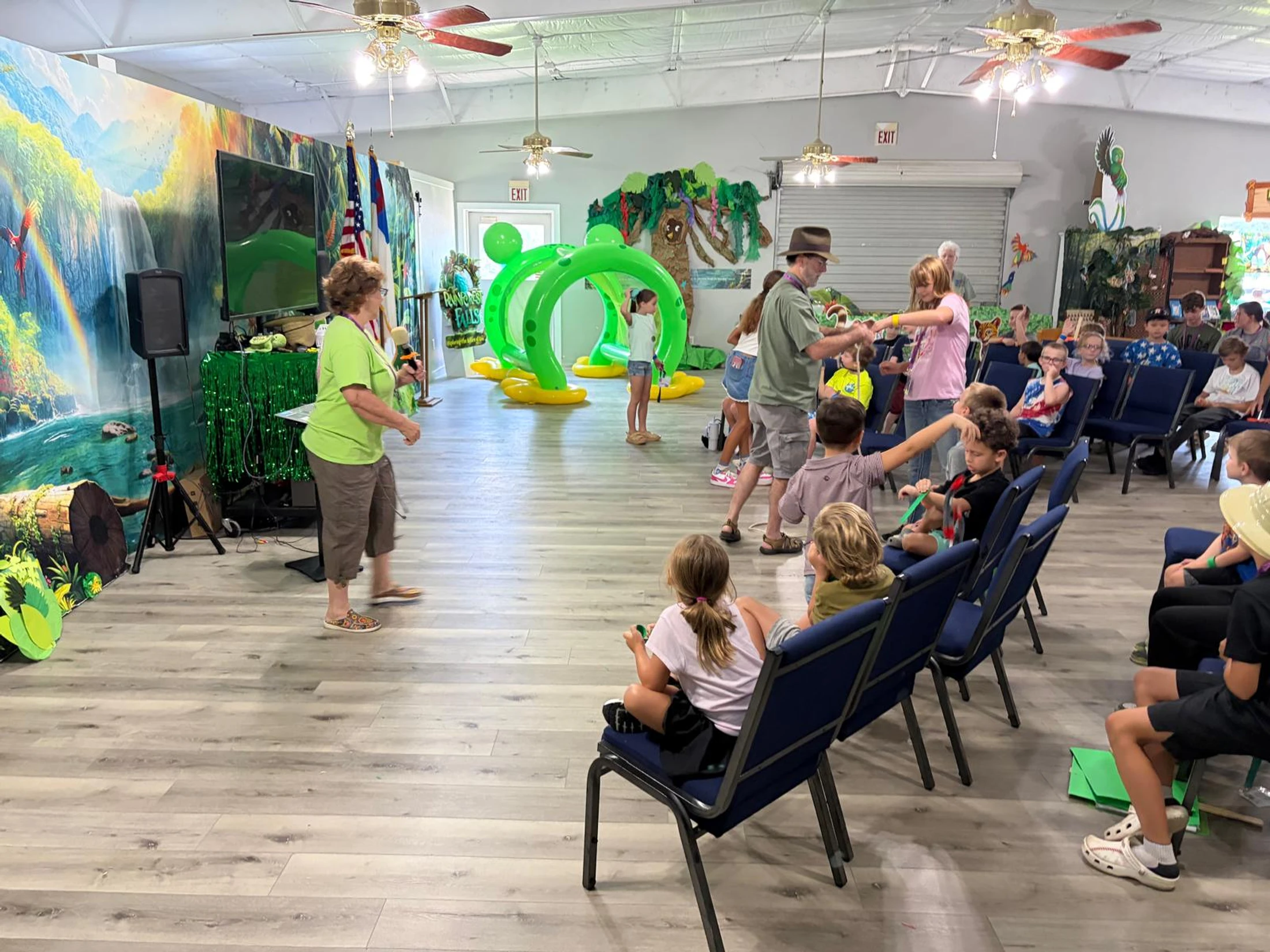 A large room decorated with a rainforest theme where children sit in chairs and participate in an activity led by several adults during an event.