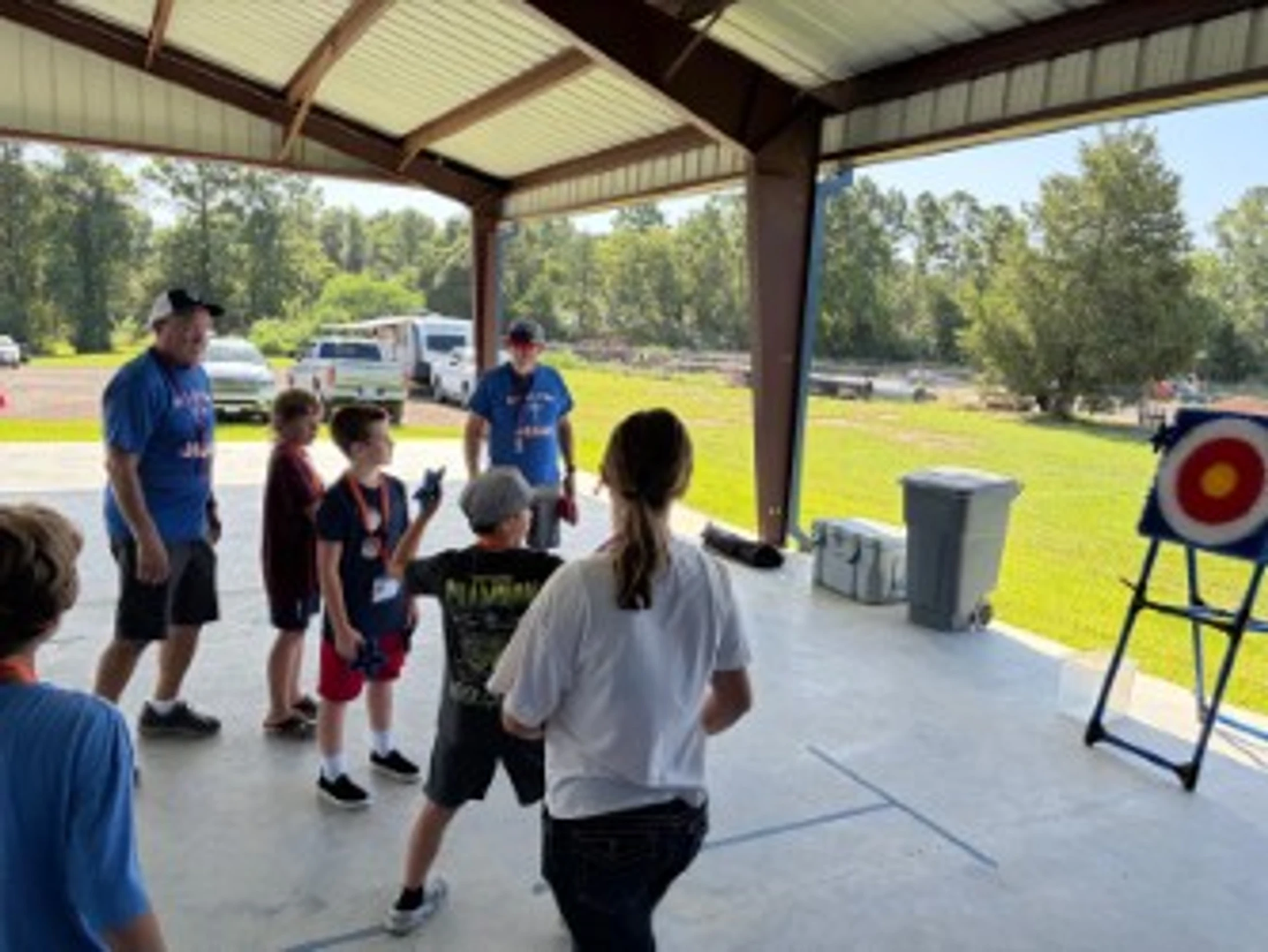 A group of children and adults under a covered pavilion, taking turns aiming at a bullseye target on a stand during a sunny day.