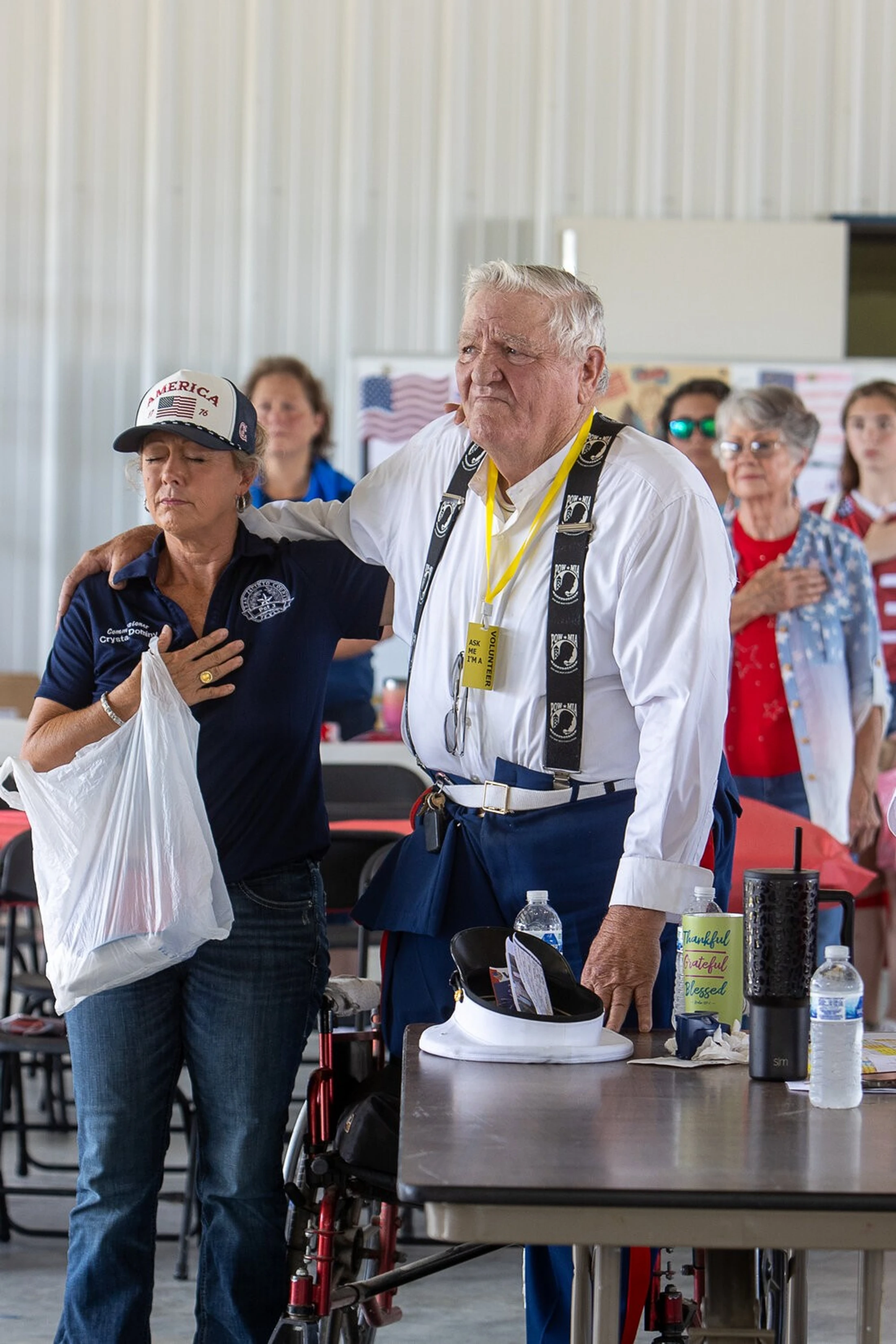 An elderly veteran with his arm around a woman as they stand with hands over their hearts during a patriotic ceremony, with others in the background.