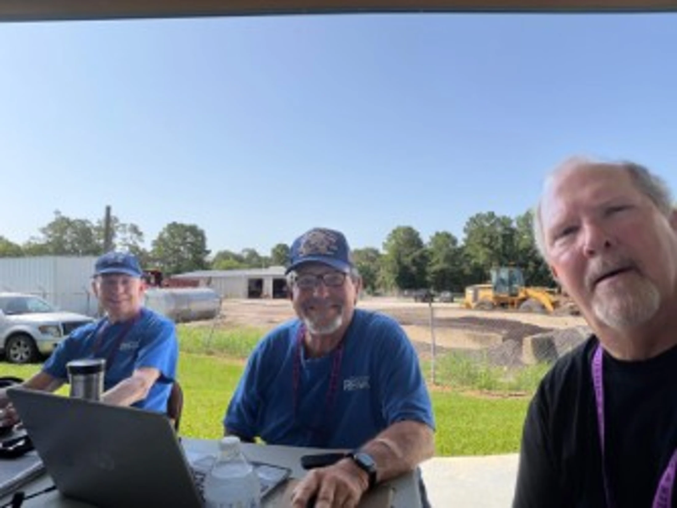 A selfie of three older men sitting at a table outdoors under a pavilion. Two wear matching blue shirts and all wear hats. A laptop is on the table.