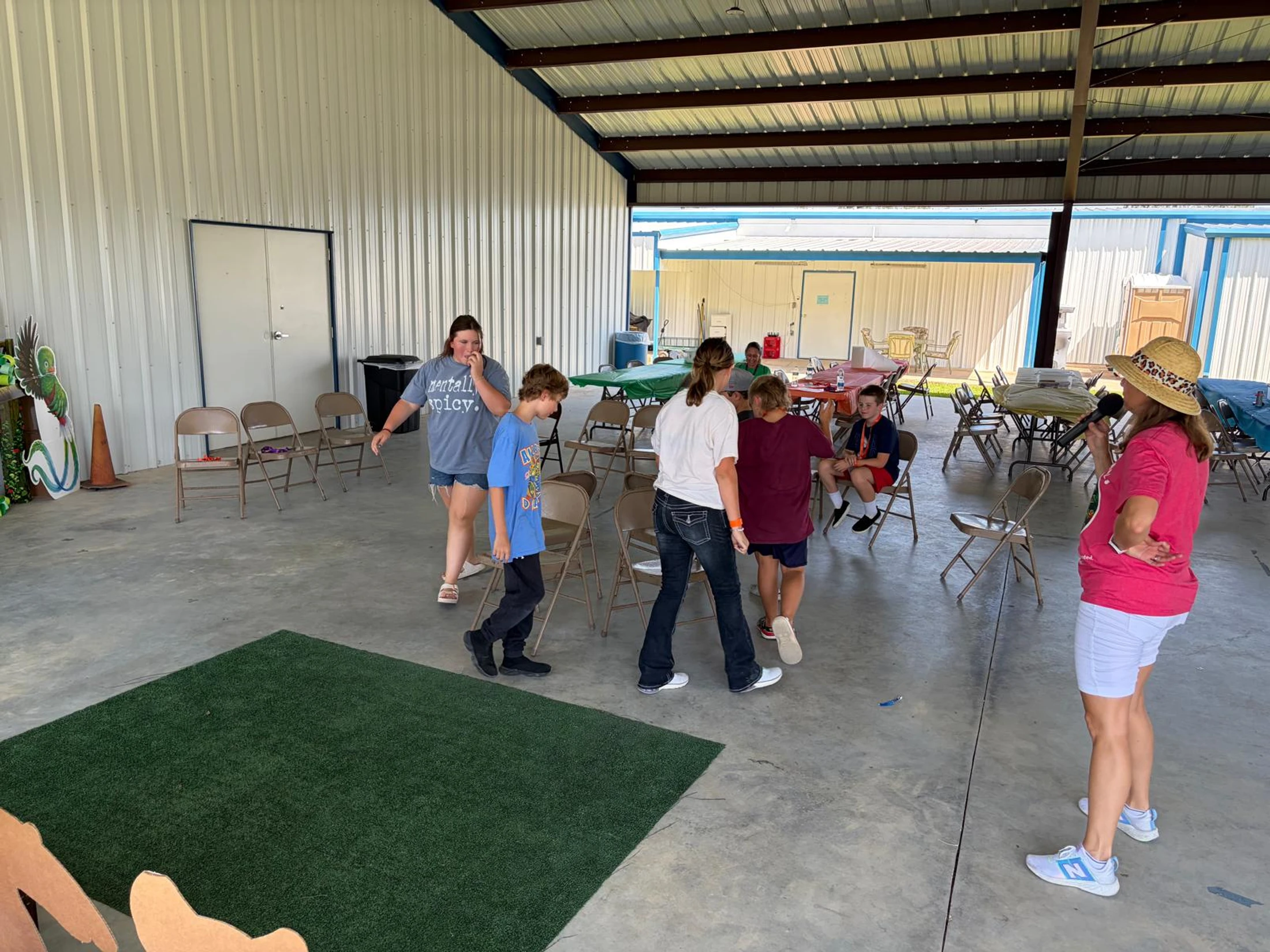 A group of children play a game like musical chairs in an open-air pavilion as an adult woman with a microphone leads the activity.