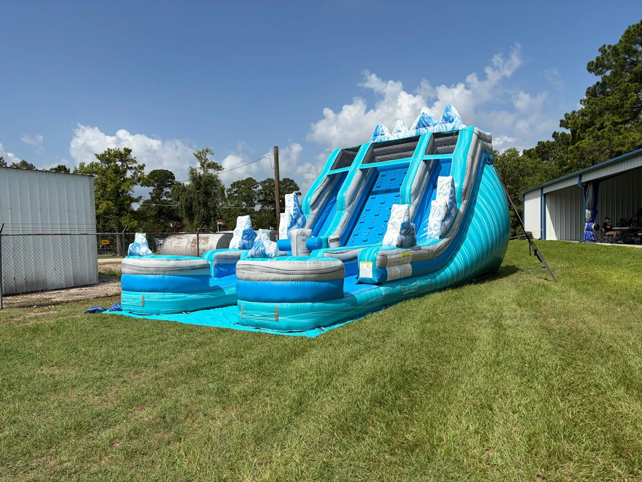 A large, blue and grey ice-themed inflatable water slide with two lanes and a climbing wall, set up on a green lawn under a partly cloudy blue sky.