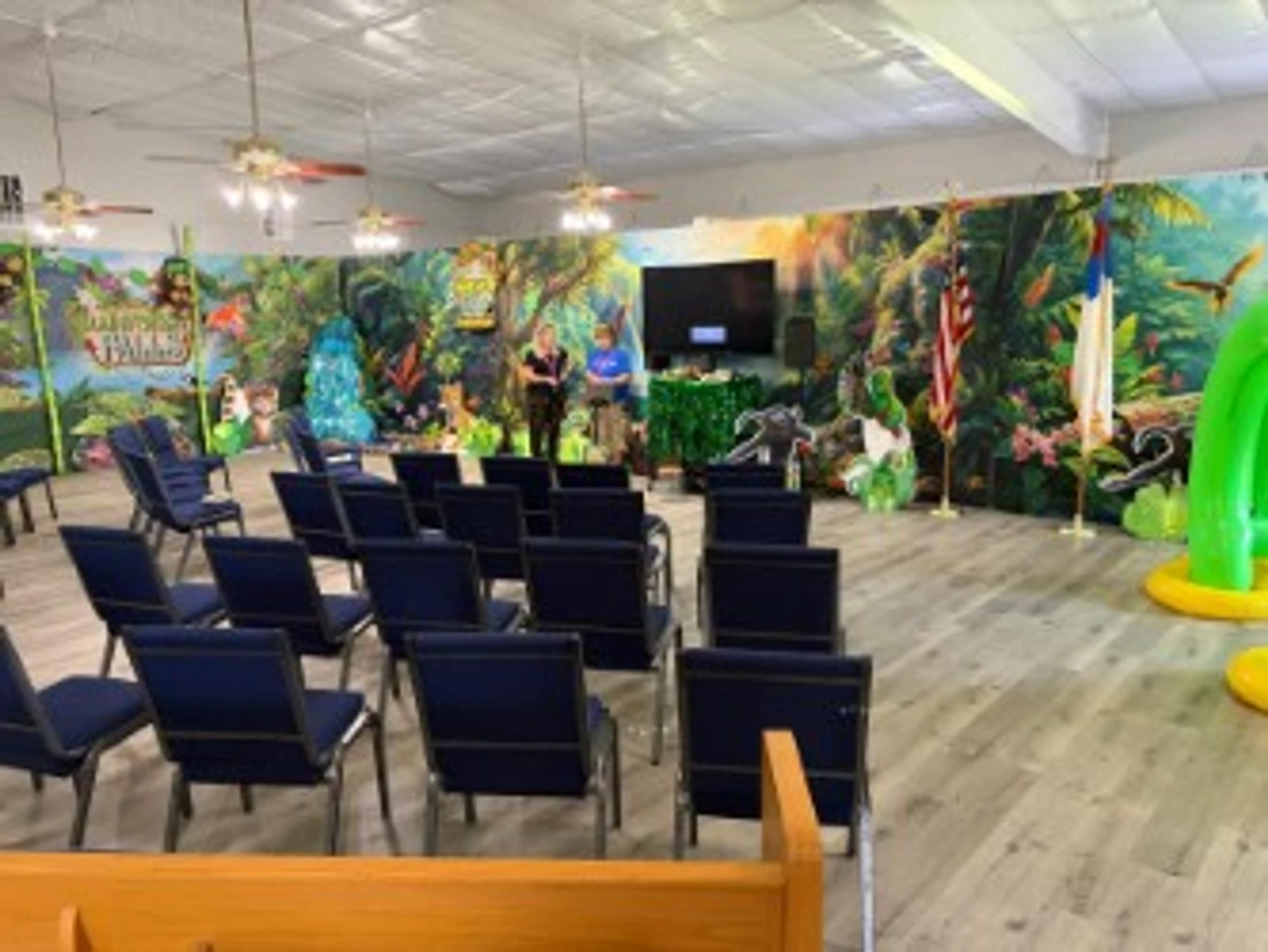 A large room set up for an event, with blue chairs facing a stage. The room is decorated with a colorful, full-wall jungle mural and inflatable decorations.