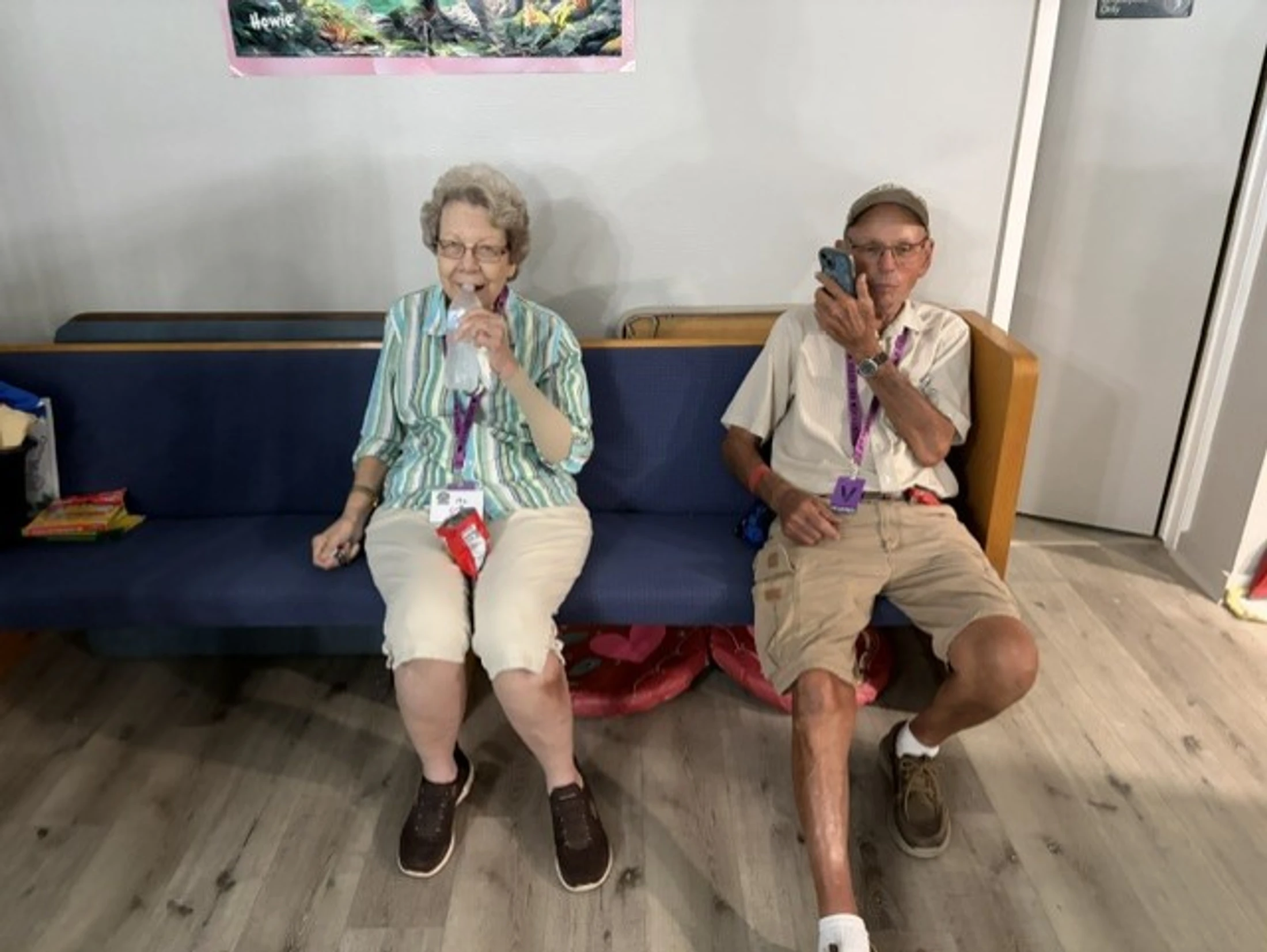 An elderly woman and man sit on a blue bench indoors. The woman smiles and drinks from a water bottle, while the man holds a phone to his ear.