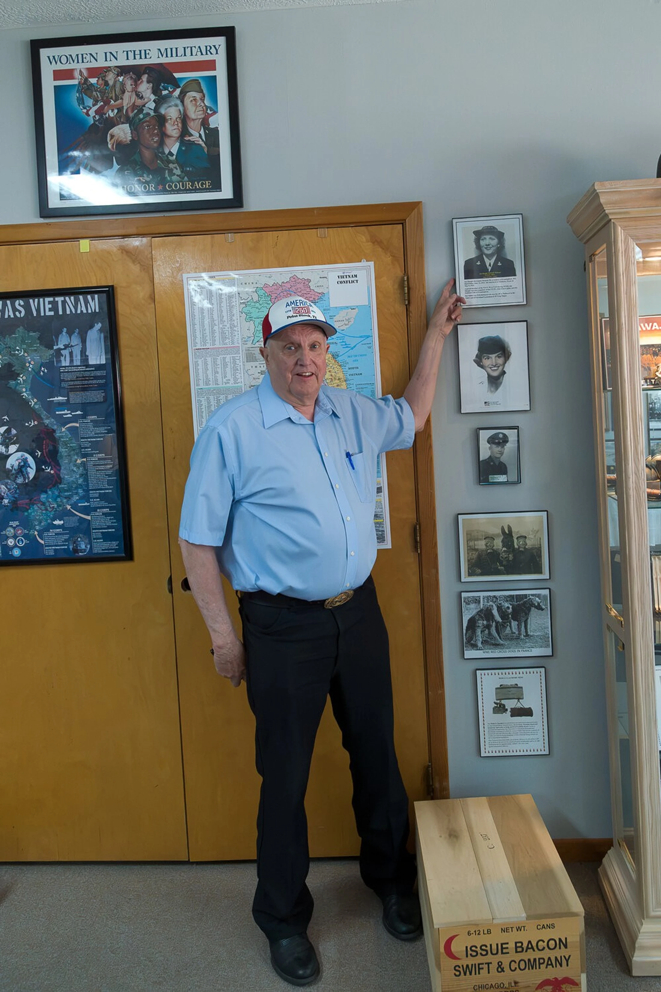 An older man in a blue shirt and baseball cap stands in a room with military memorabilia, pointing to a wall of framed historical photos.