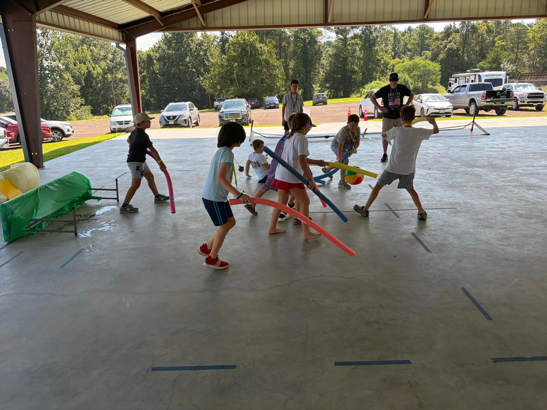 A group of kids at a summer camp playing an energetic game with colorful pool noodles and a beach ball under a large outdoor pavilion.
