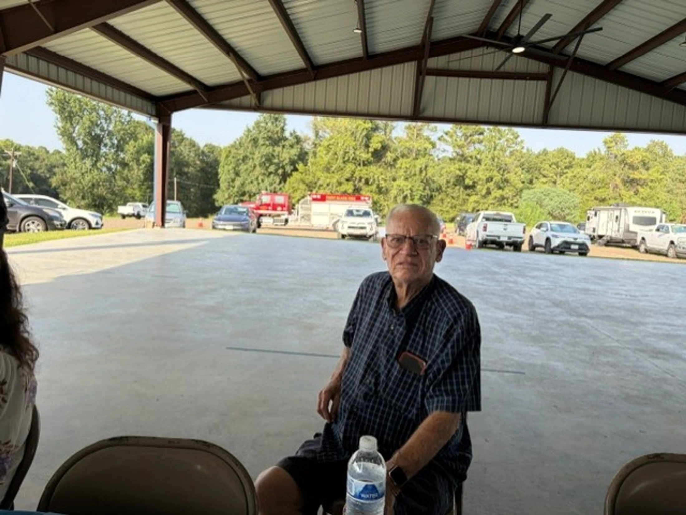 An elderly man in a plaid shirt and glasses sits under a pavilion, looking at the camera. A parking lot with cars and a fire truck is visible behind him.
