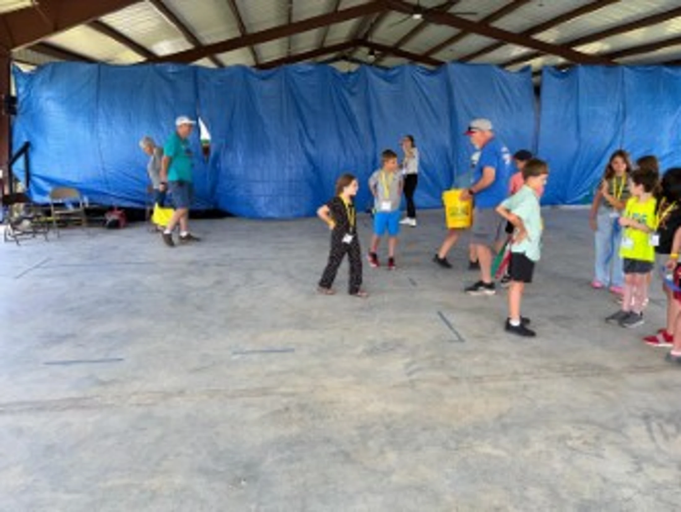 A group of children and a few adults gather on a concrete floor inside a large pavilion with a blue tarp backdrop, seemingly playing an organized game.