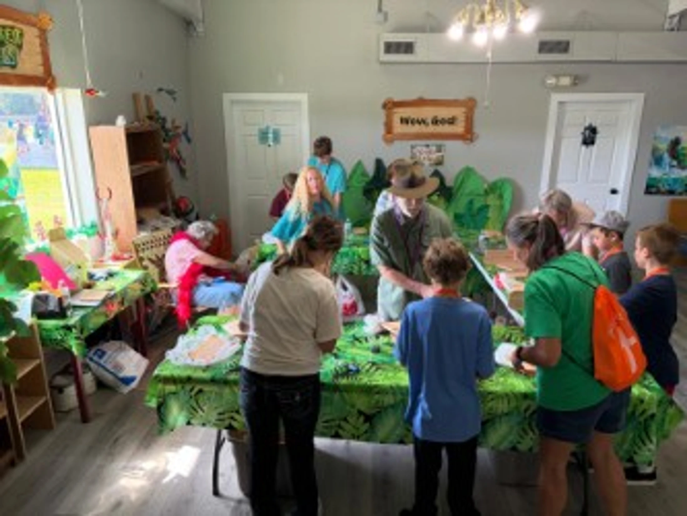A group of children and adults gathered around tables with leaf-print tablecloths, participating in a nature-themed craft workshop led by a man in a safari hat.
