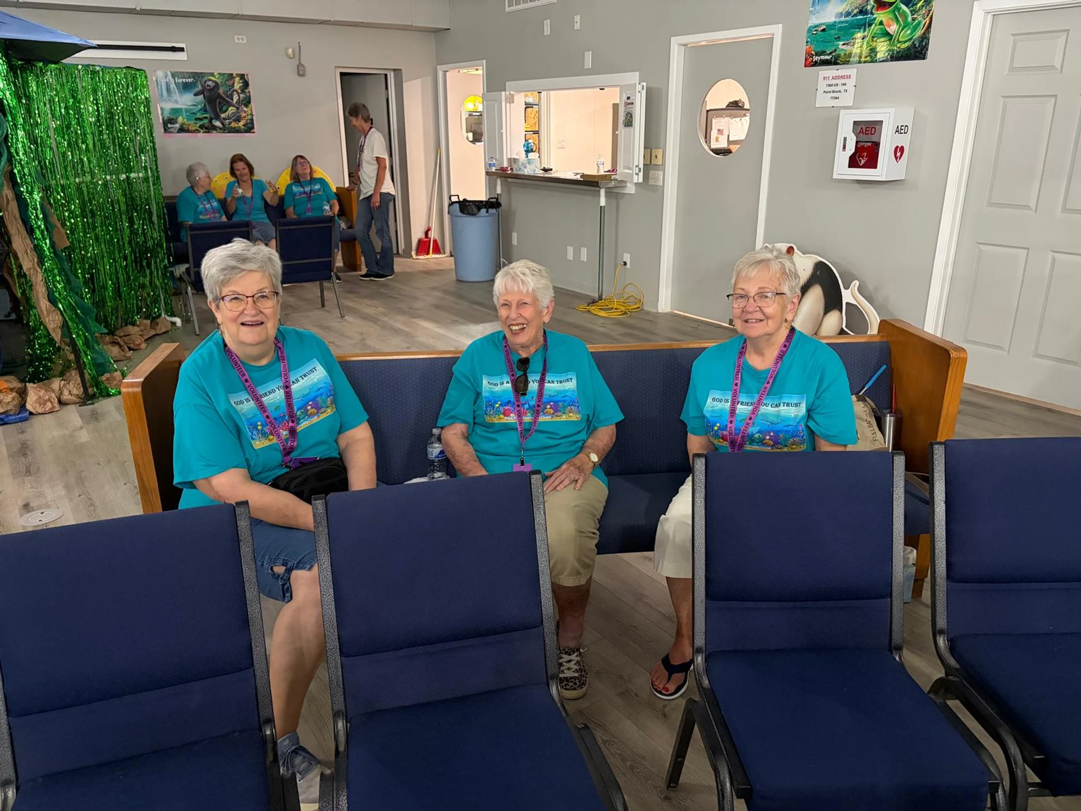 Three smiling older women in matching turquoise volunteer t-shirts sit together on a bench in a community hall during an event.