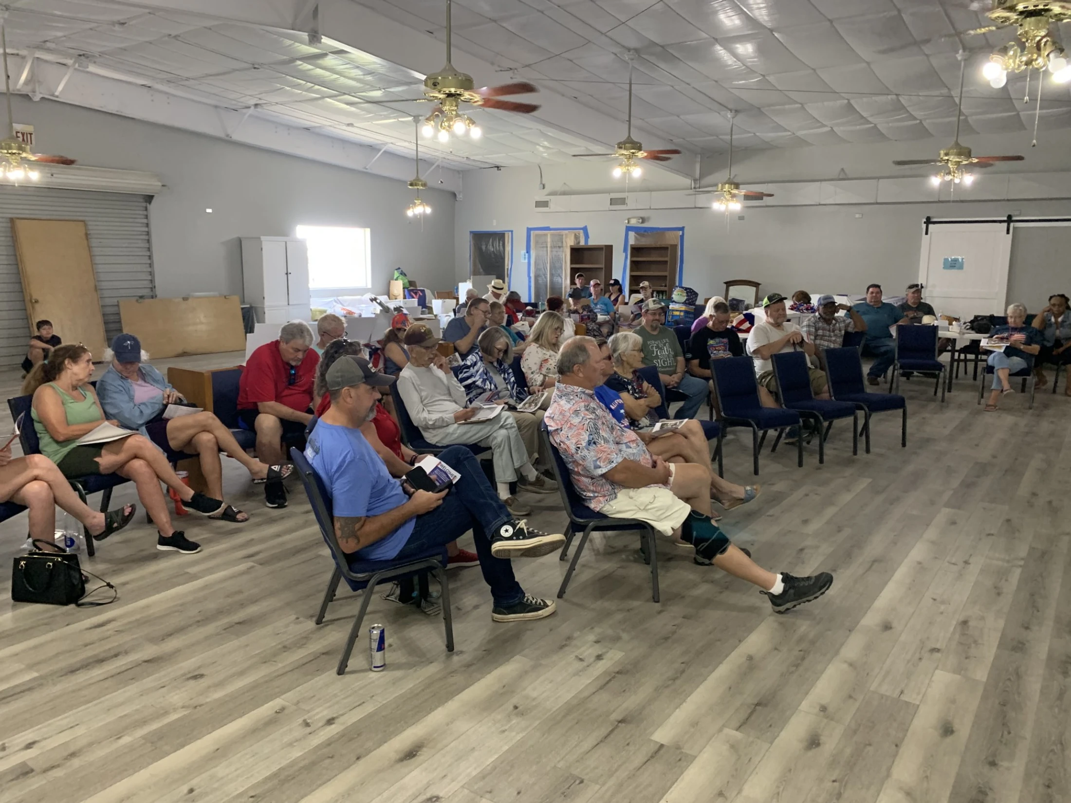 A crowd of people seated in a large, bright room with a wood-look floor, listening during a community meeting or presentation.