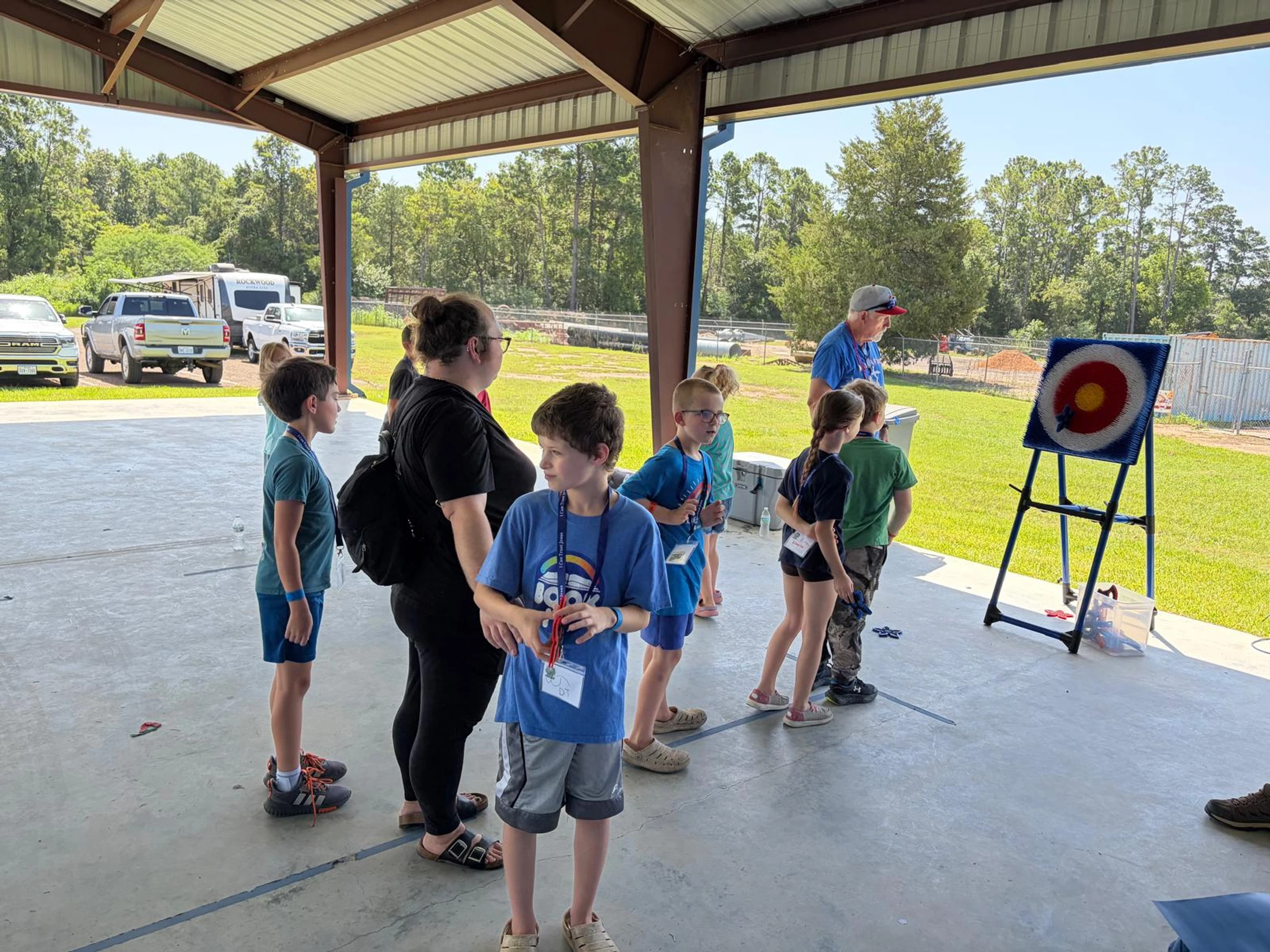 A group of children and a few adults gather under a pavilion on a sunny day, taking turns with a kid-safe dart and target game.