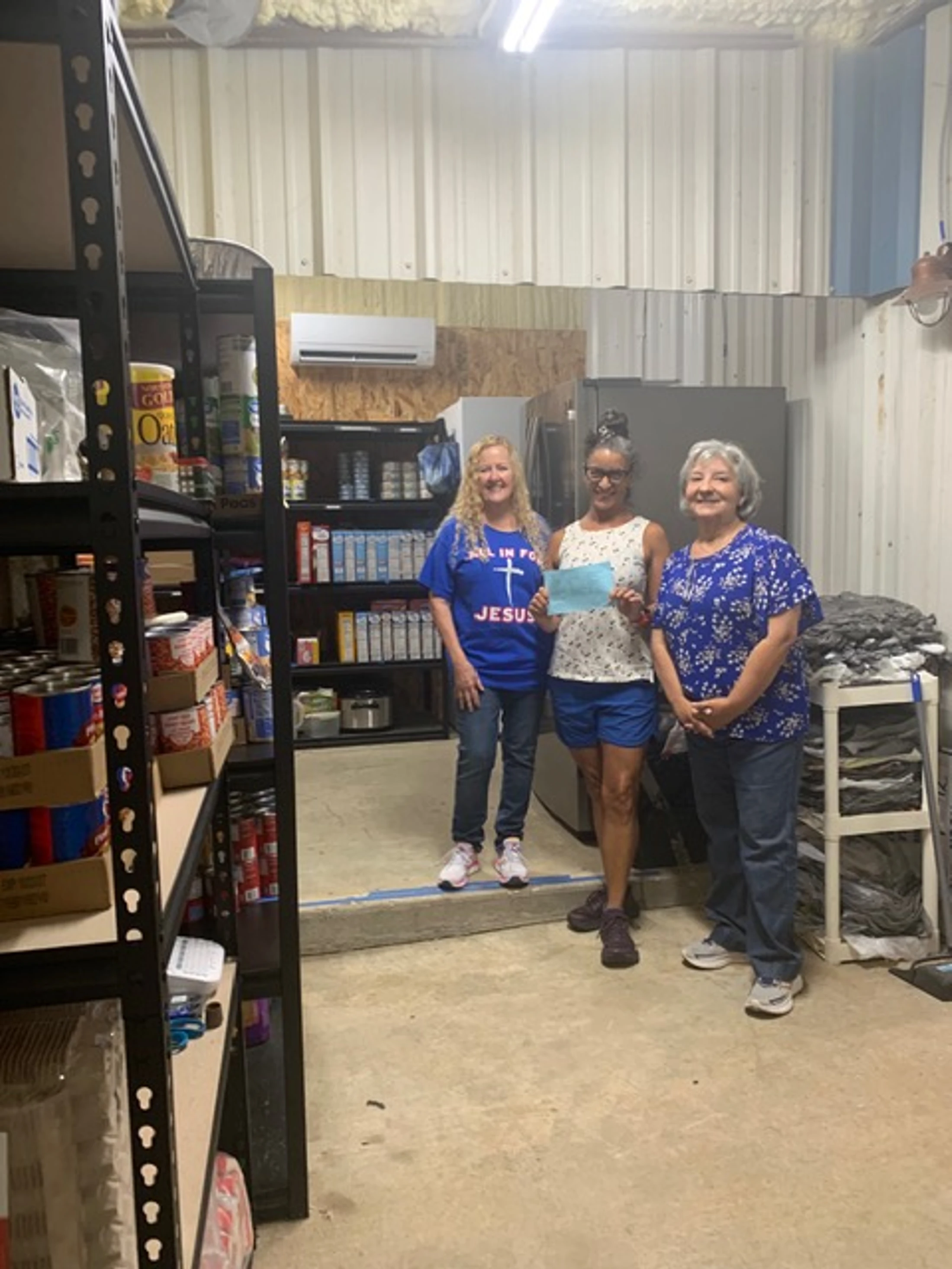 Three women smile while standing in a storage room filled with shelves of canned goods and food. The woman in the center holds a light blue envelope.