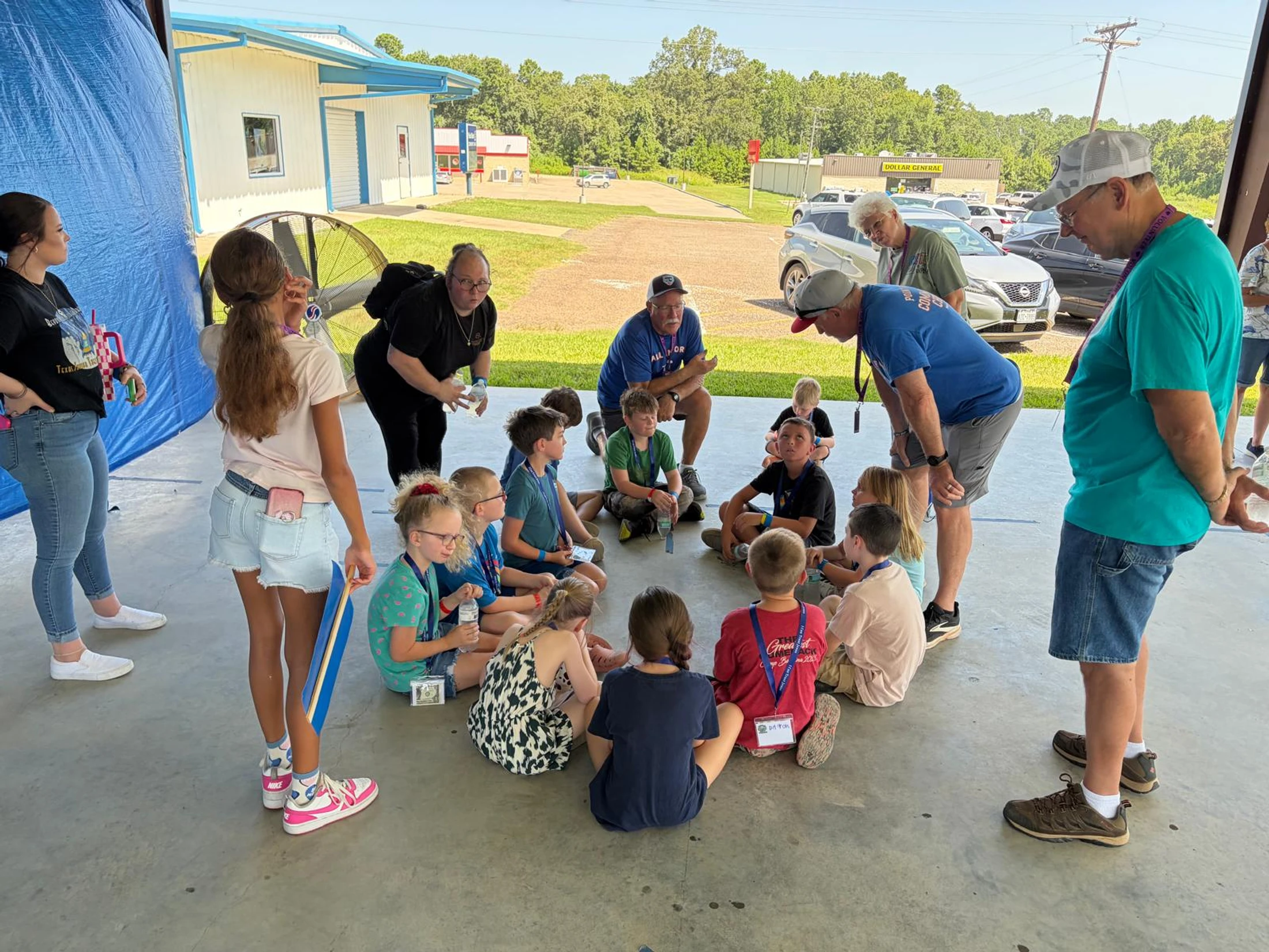 A group of children sits in a circle on a concrete floor, listening as several adult volunteers talk with them during an outdoor event under a covered area.
