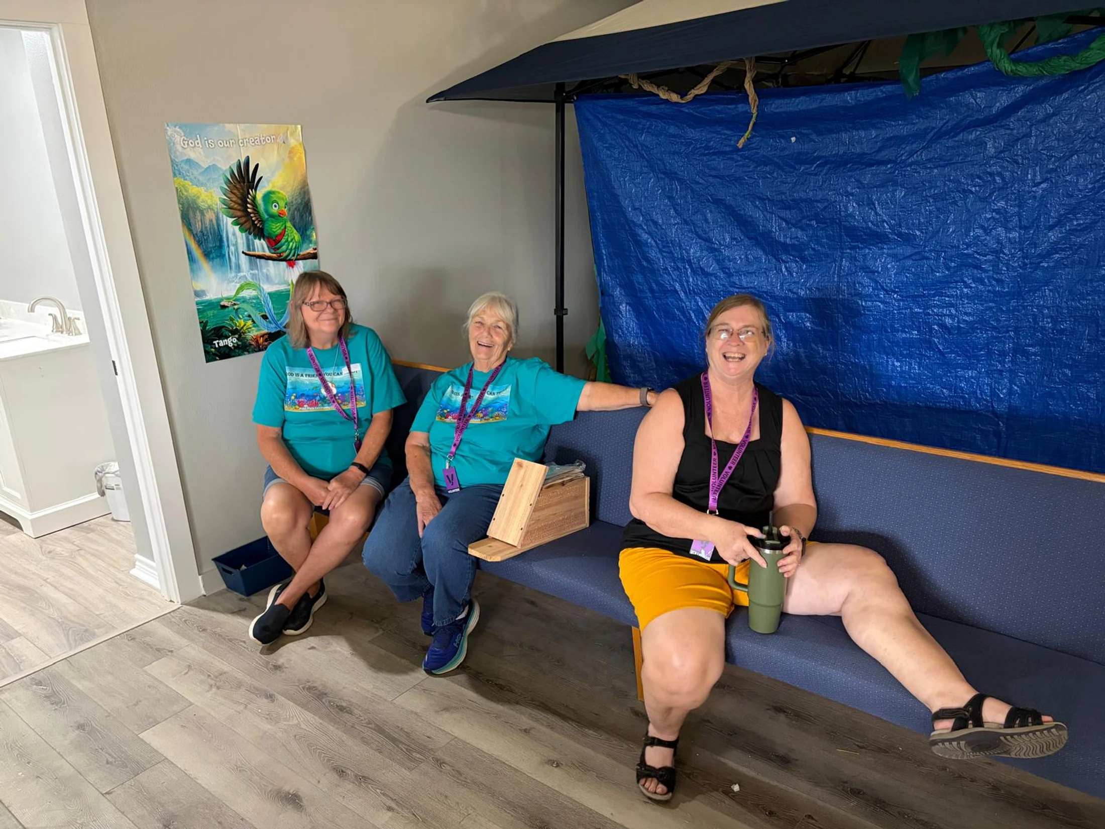 Three smiling women sit together on a blue bench indoors. Two wear matching turquoise volunteer t-shirts, and all three wear purple volunteer lanyards.