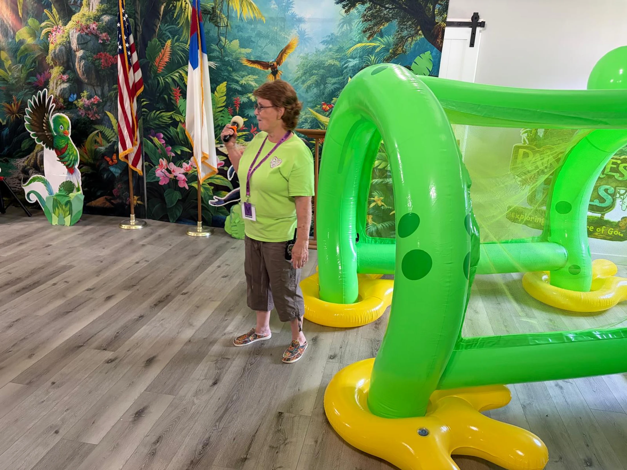 A woman in a green shirt holds a microphone, standing next to a large green inflatable structure in a room decorated with a jungle mural.