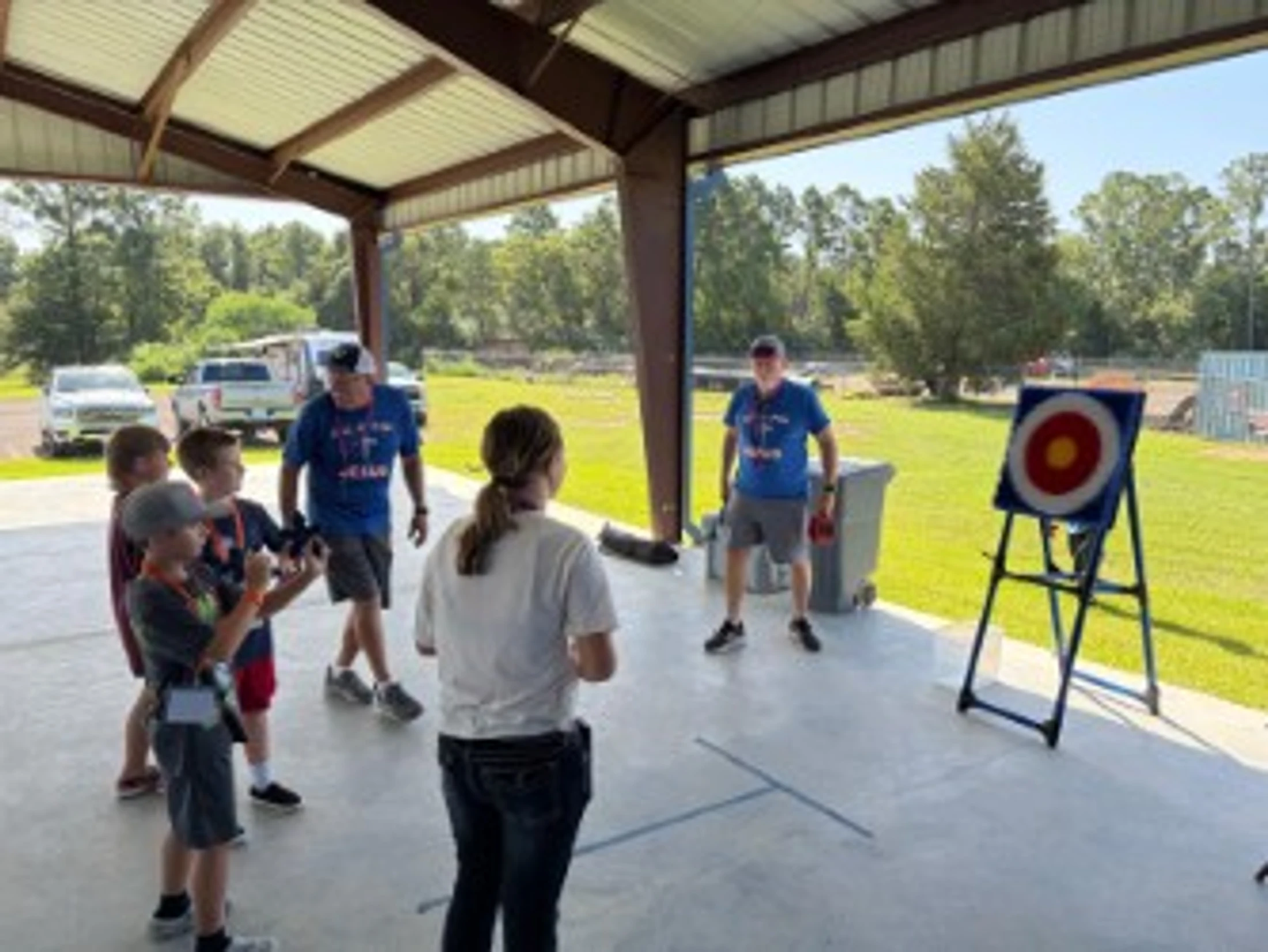 A group of children and two adult instructors learning archery under a pavilion. One child is aiming a bow at a target on a stand.