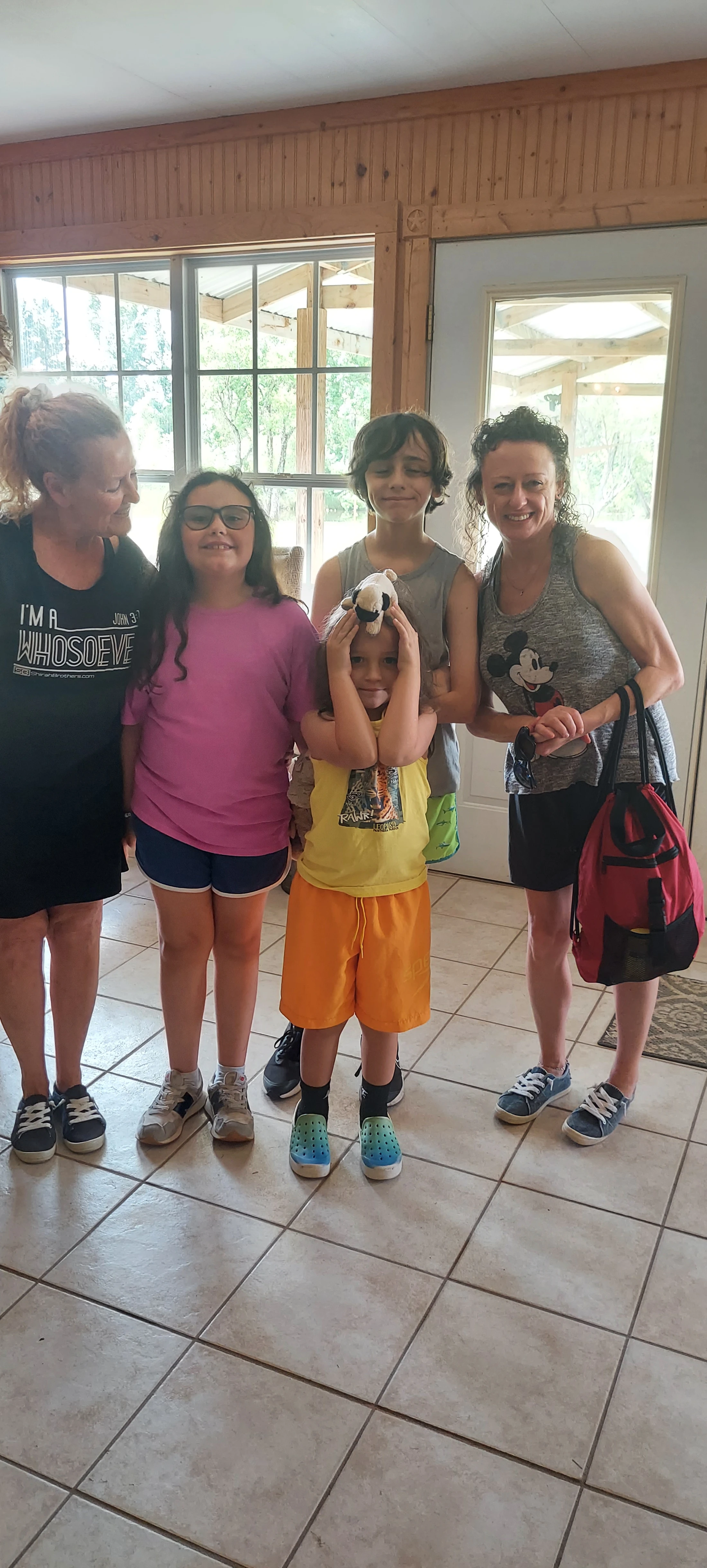 A group of two women and three children posing for a photo on a tiled floor. A small child in the center makes a silly face with a stuffed pug on their head.