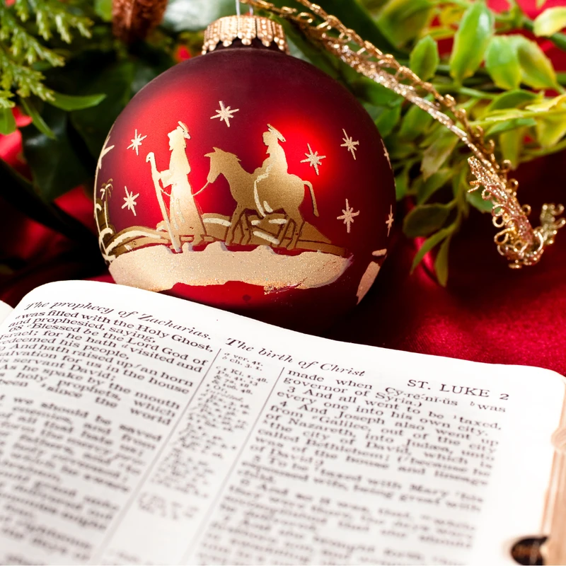 A red Christmas ornament with a golden nativity scene rests on an open Bible, turned to the story of Christ's birth, surrounded by festive greenery.