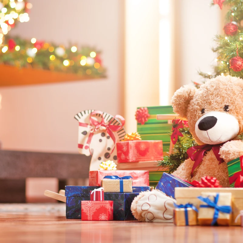 A large teddy bear sits among a pile of wrapped presents under a lit Christmas tree, with a cozy fireplace glowing in the background.