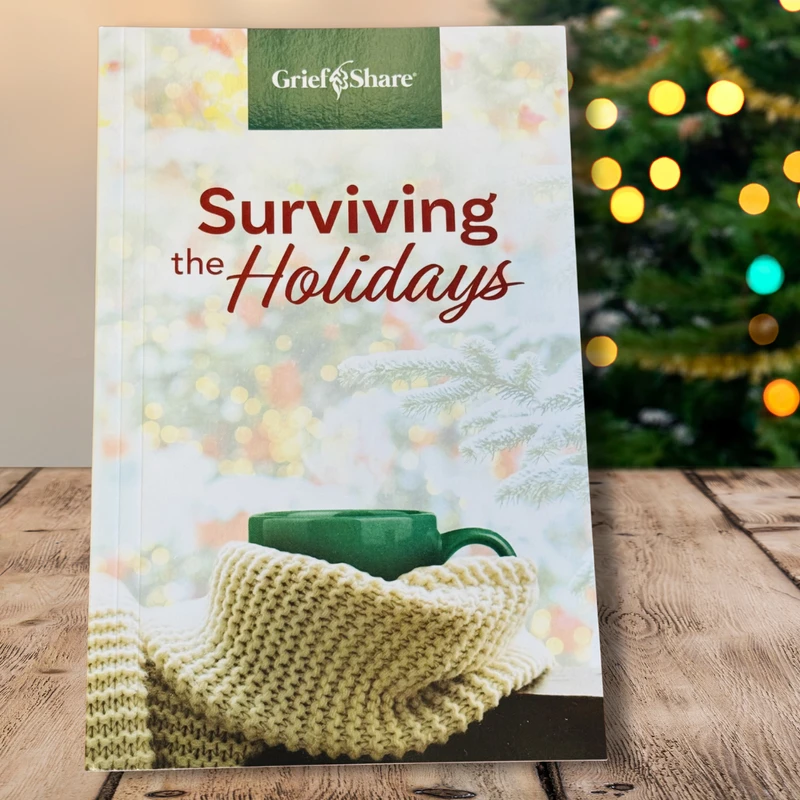 The 'Surviving the Holidays' booklet from GriefShare rests on a wooden table with a softly lit Christmas tree in the background.
